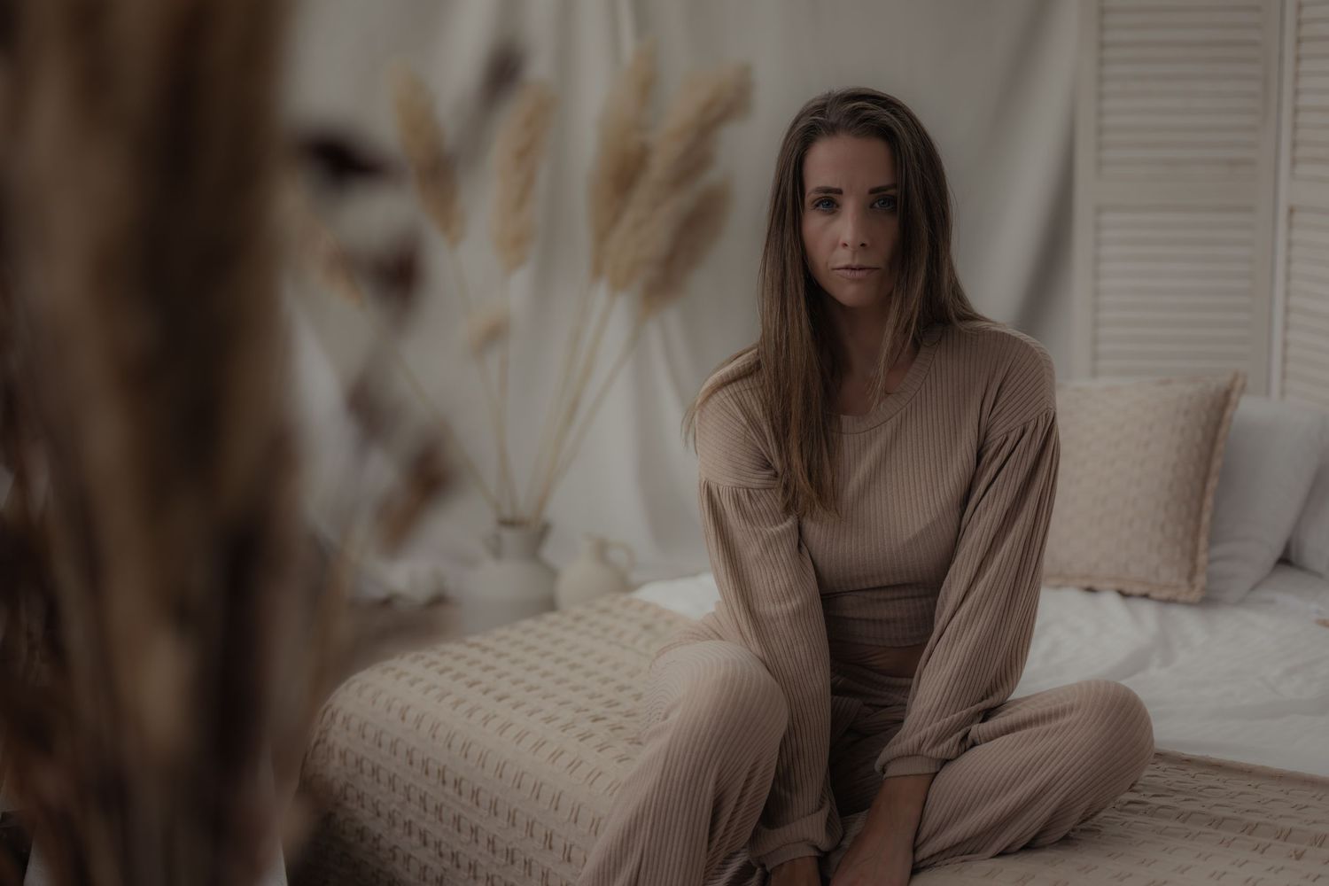 Woman in beige loungewear sitting cross-legged on a bed in a cozy, neutral-toned bedroom with dried pampas grass.