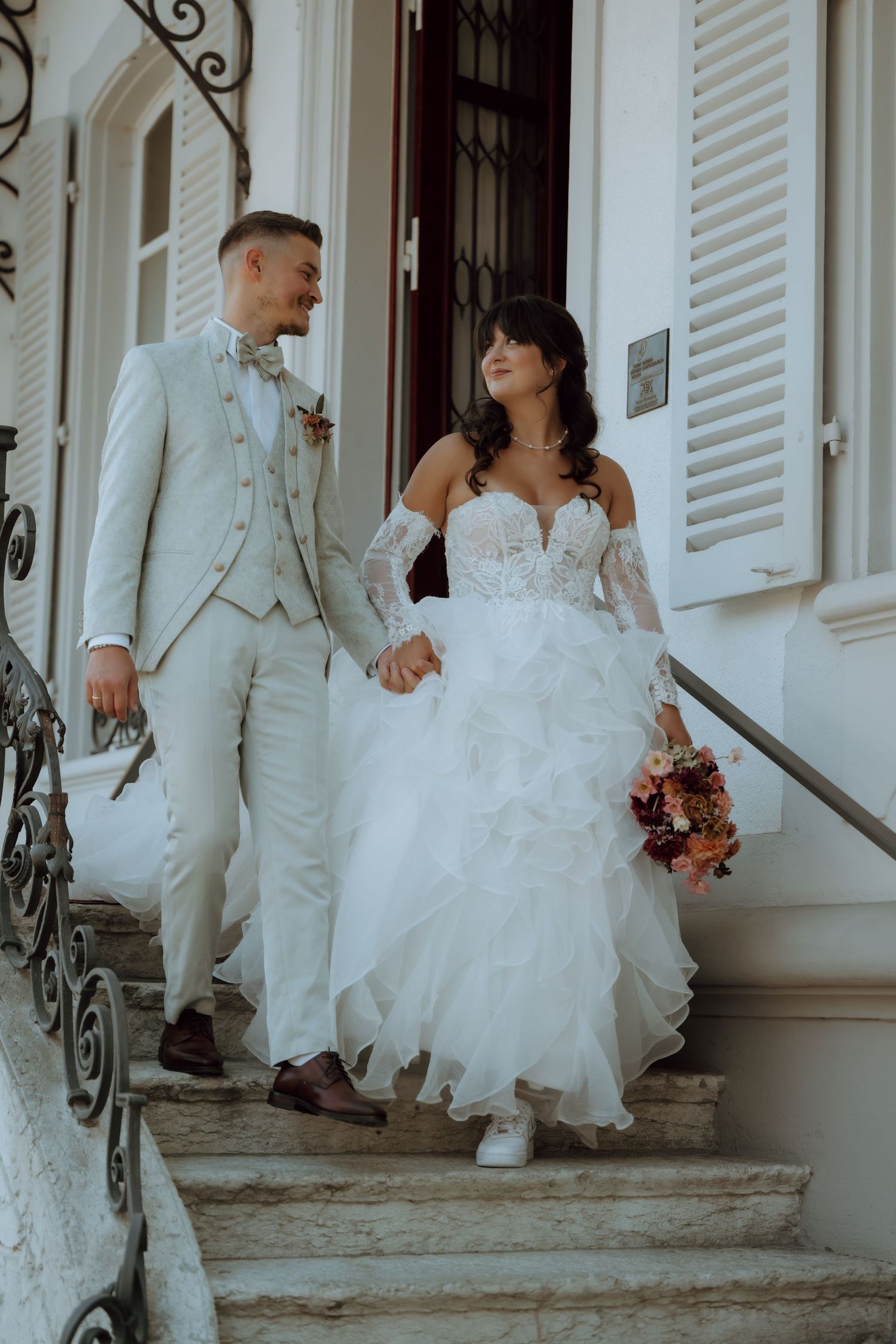 Bride and groom holding hands walking down stone steps, she in ruffled white gown, he in light gray suit.