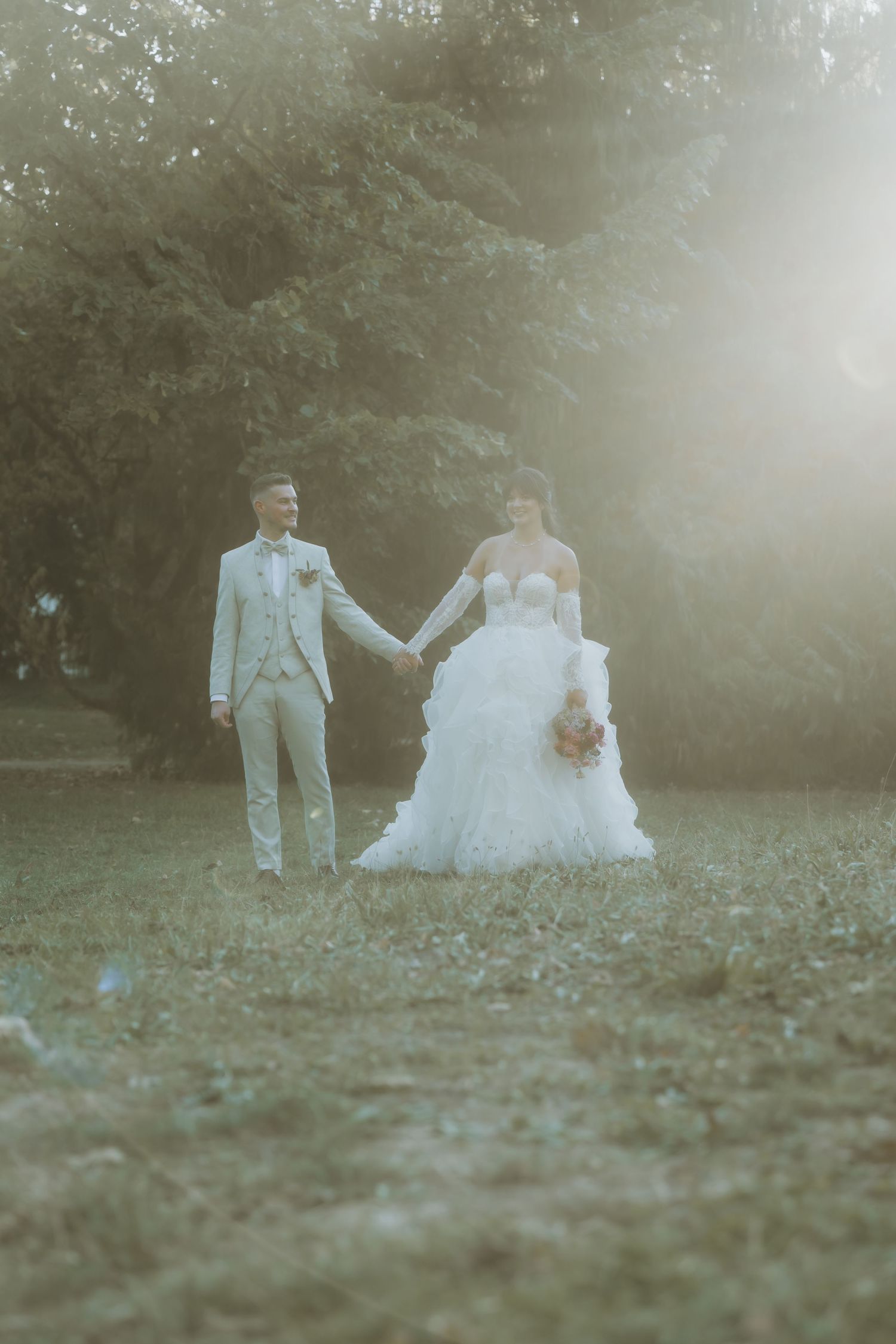 Bride and groom holding hands walking through sunlit garden, bride in ruffled white gown, groom in light beige suit.
