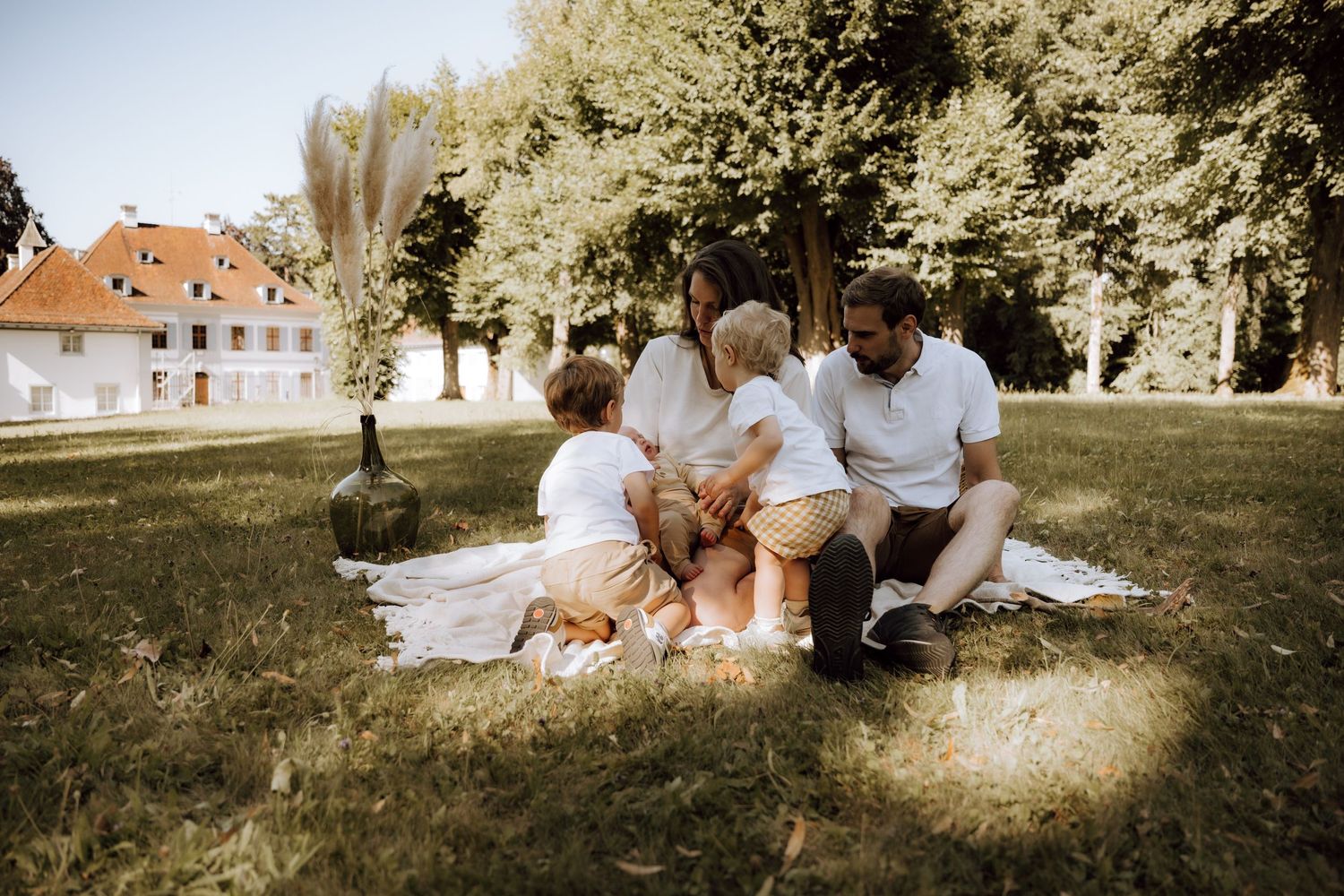 Family of four enjoying a sunny picnic on a blanket in a lush green park surrounded by trees.