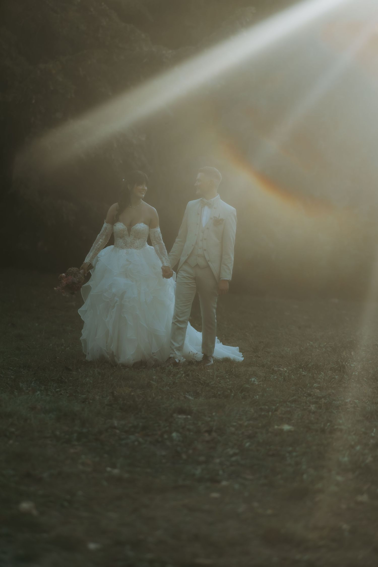 Bride and groom holding hands walking through sunlit field, bride in ruffled white gown, groom in tan suit.