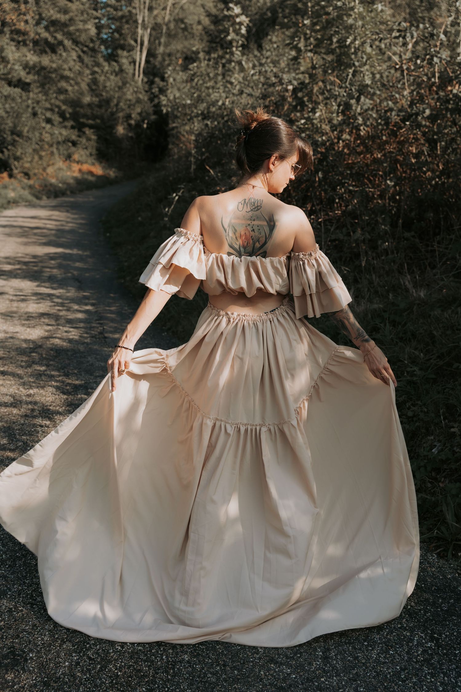 Woman in elegant champagne off-shoulder gown with back tattoo walks along a forest path.