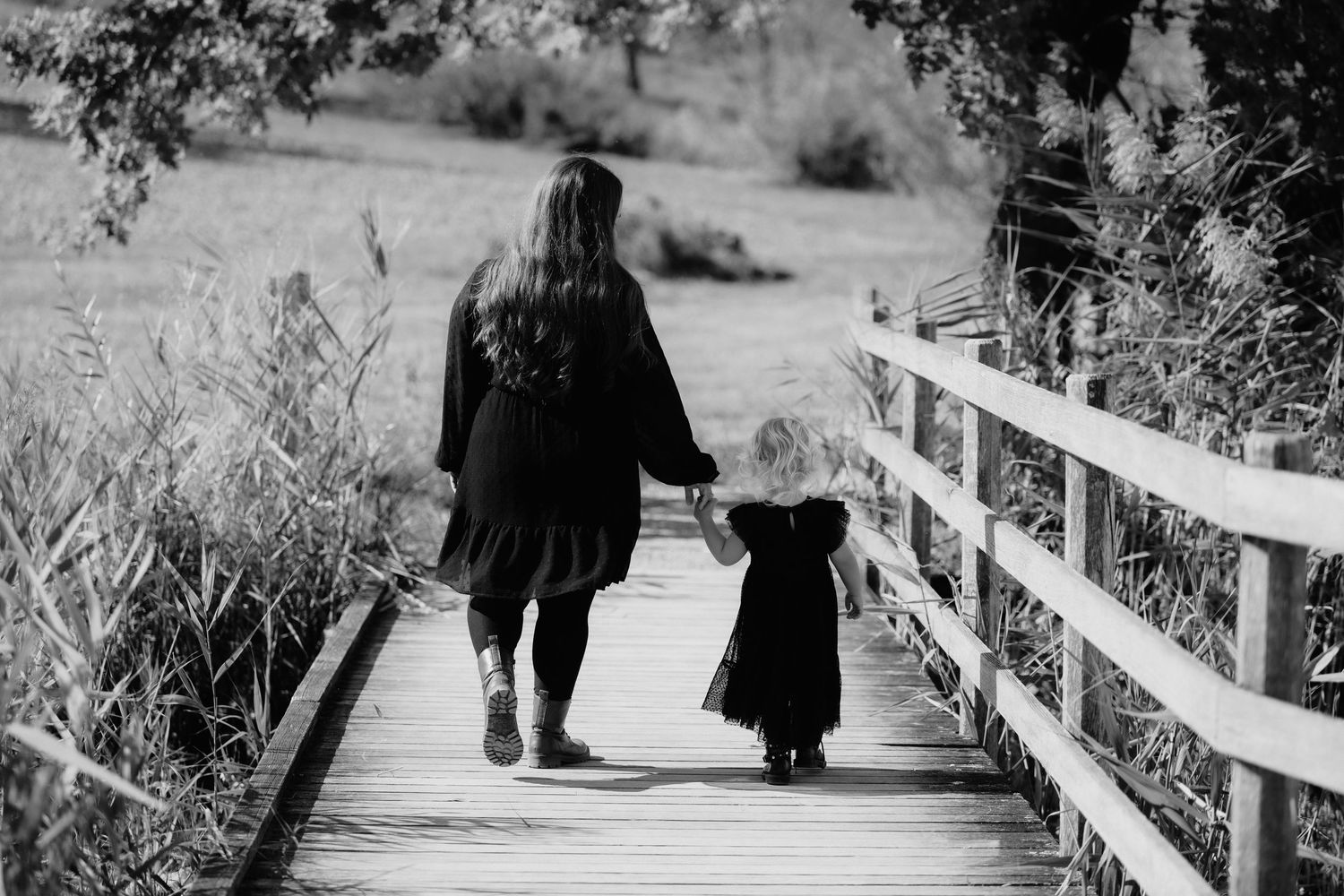 Black and white photo of a mother and young daughter holding hands while walking on a wooden bridge path.