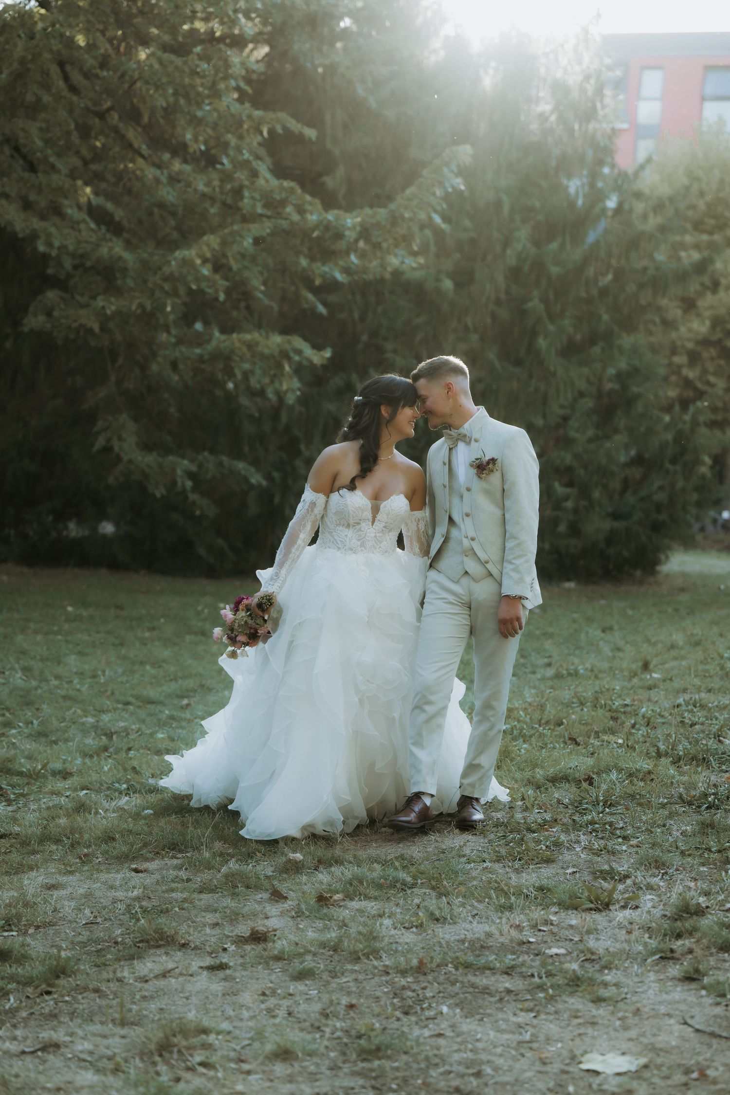 Bride and groom share a romantic moment in a sunlit garden, she in a flowing white gown, he in a light gray suit.