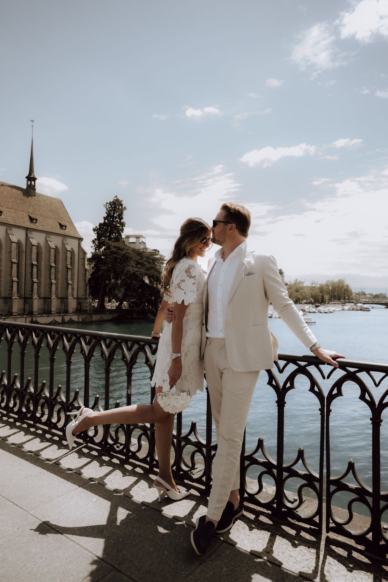 Newlyweds in white wedding outfits leaning on ornate bridge railing by a river in Zurich, Switzerland.