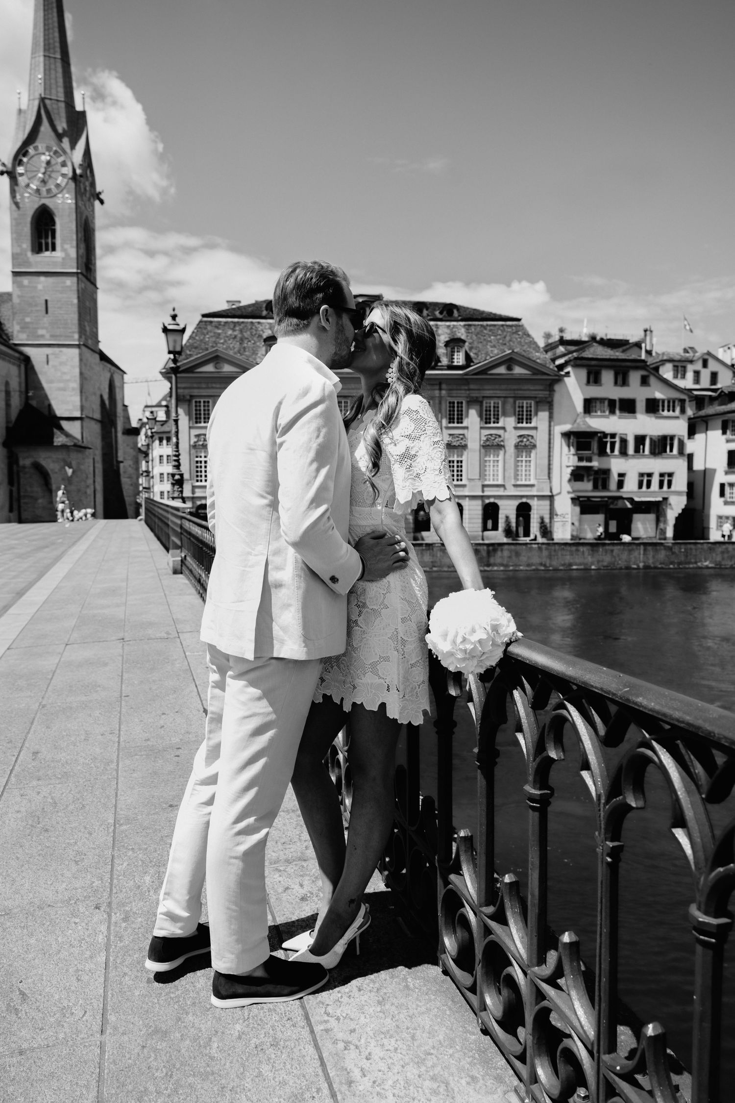 Black and white photo of newlyweds kissing on Zurich bridge, bride holding bouquet, historic church and river visible behind them.