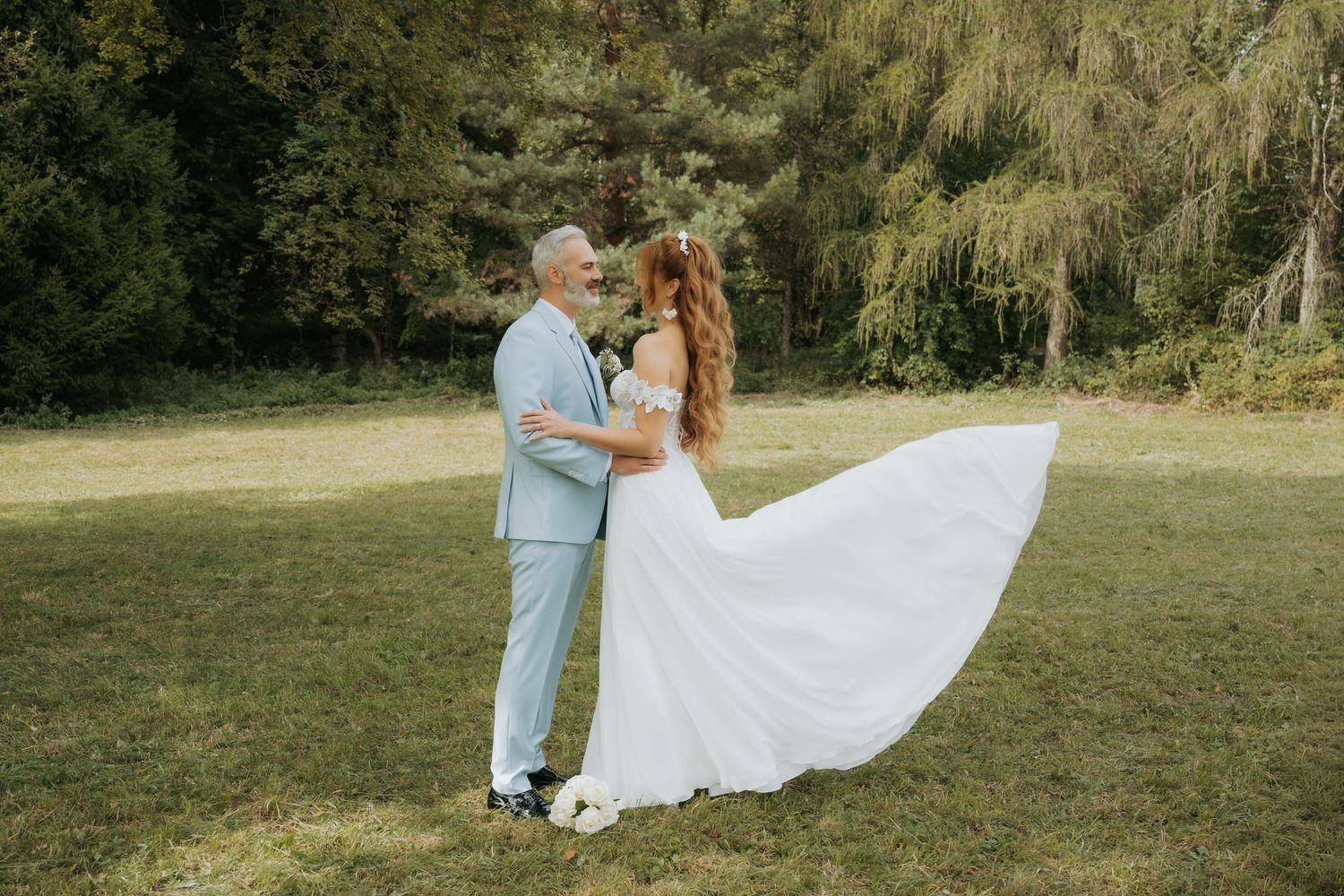 Bride in flowing white gown and groom in light blue suit share romantic dance on lush green lawn outdoors.