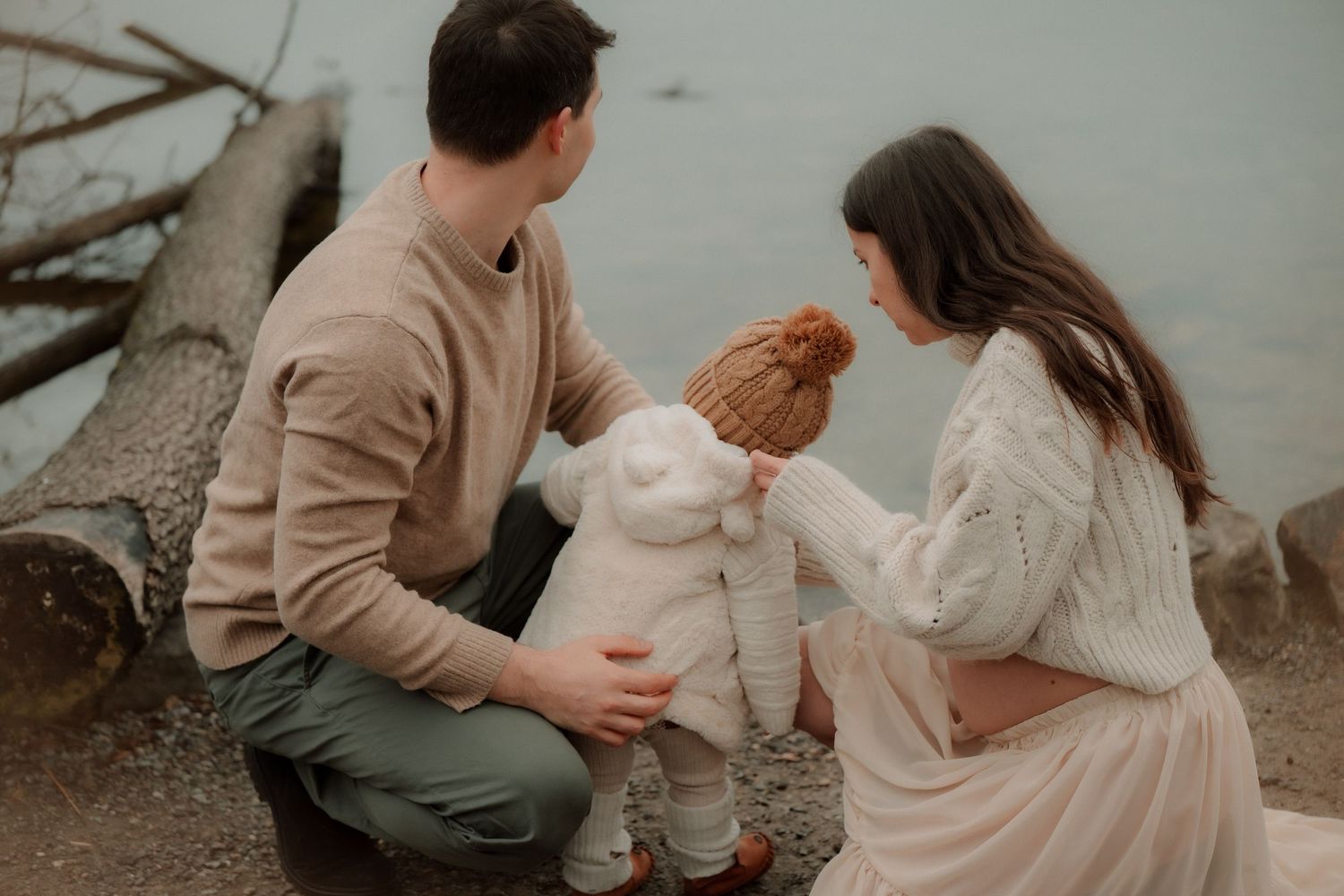 A young family with a toddler in a red hat shares a tender moment by a lakeside with driftwood.