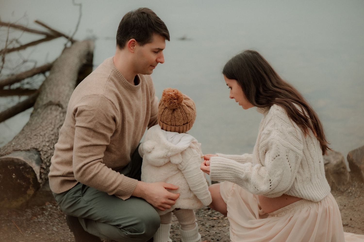 A young family with a redheaded toddler sits together on a rocky beach, sharing a tender moment near driftwood.