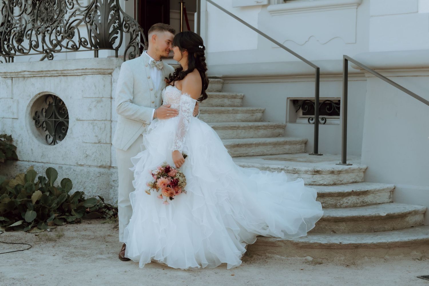 A bride in a flowing white off-shoulder gown and groom in a beige suit share a romantic moment on stone steps.