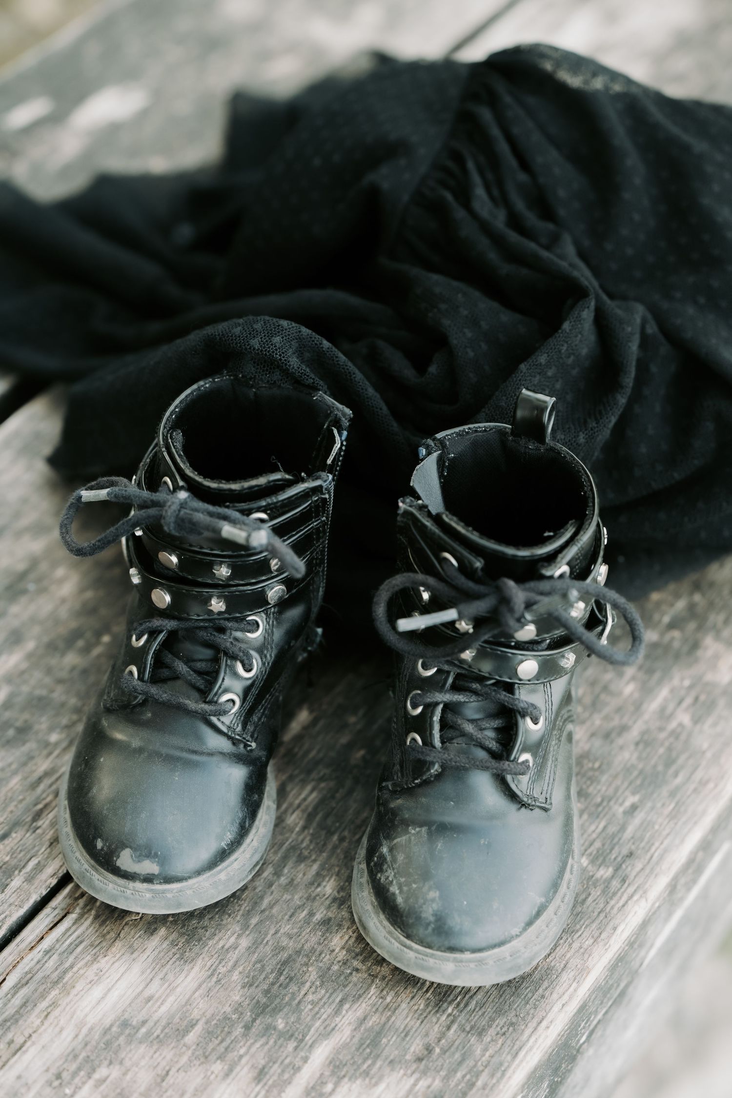 Worn black leather lace-up boots with silver studs on wooden surface beside black fabric.