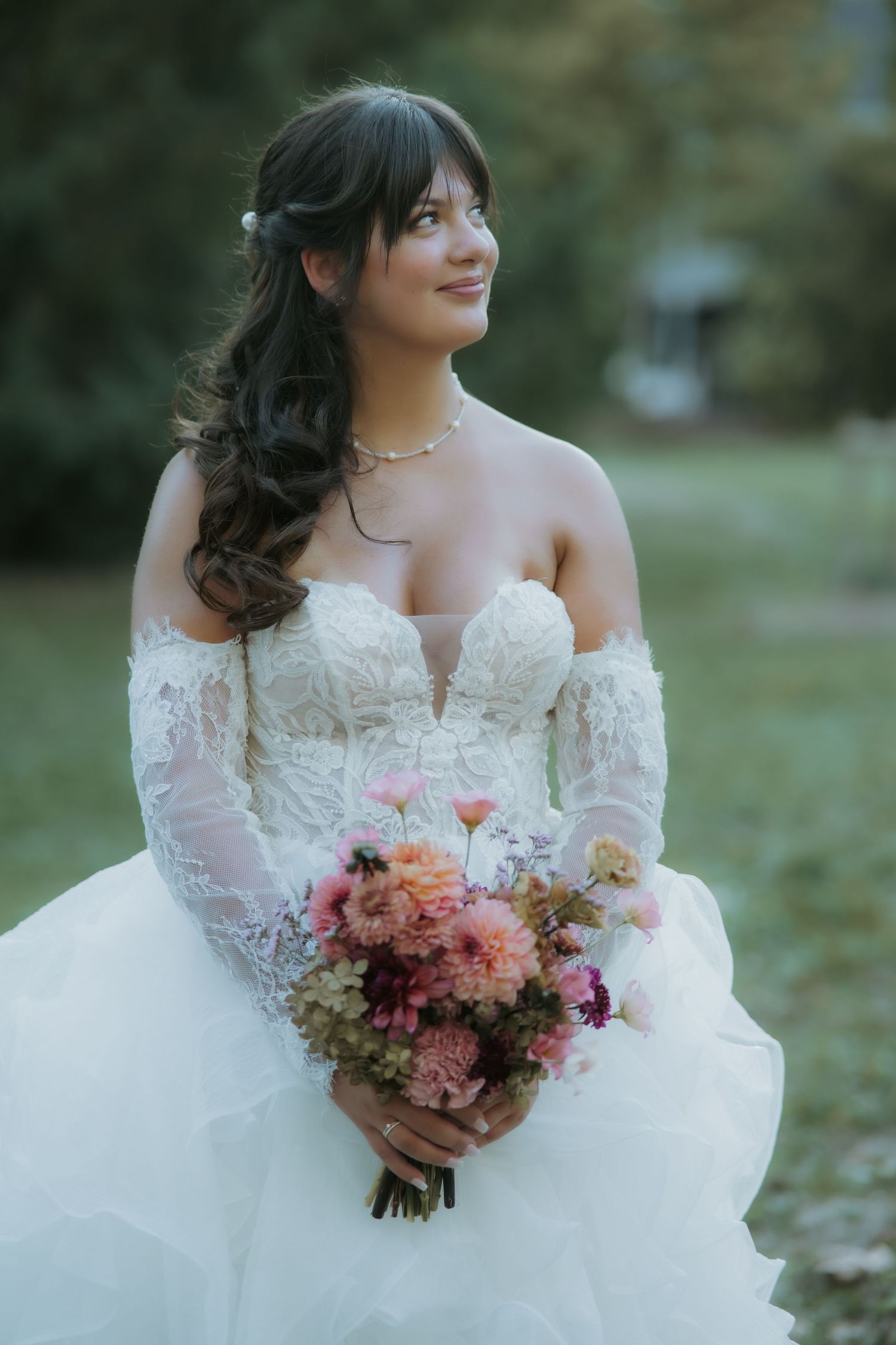 Smiling bride in off-shoulder lace wedding gown holds colorful pink bouquet outdoors among green trees.