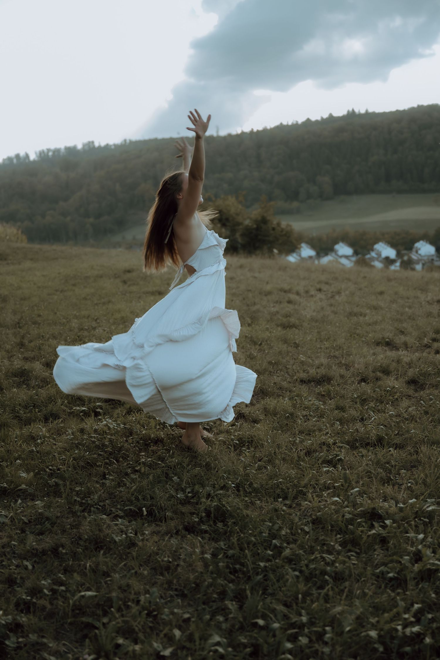 Woman in flowing white dress dancing freely in a green meadow with forested hills and dramatic cloudy sky.