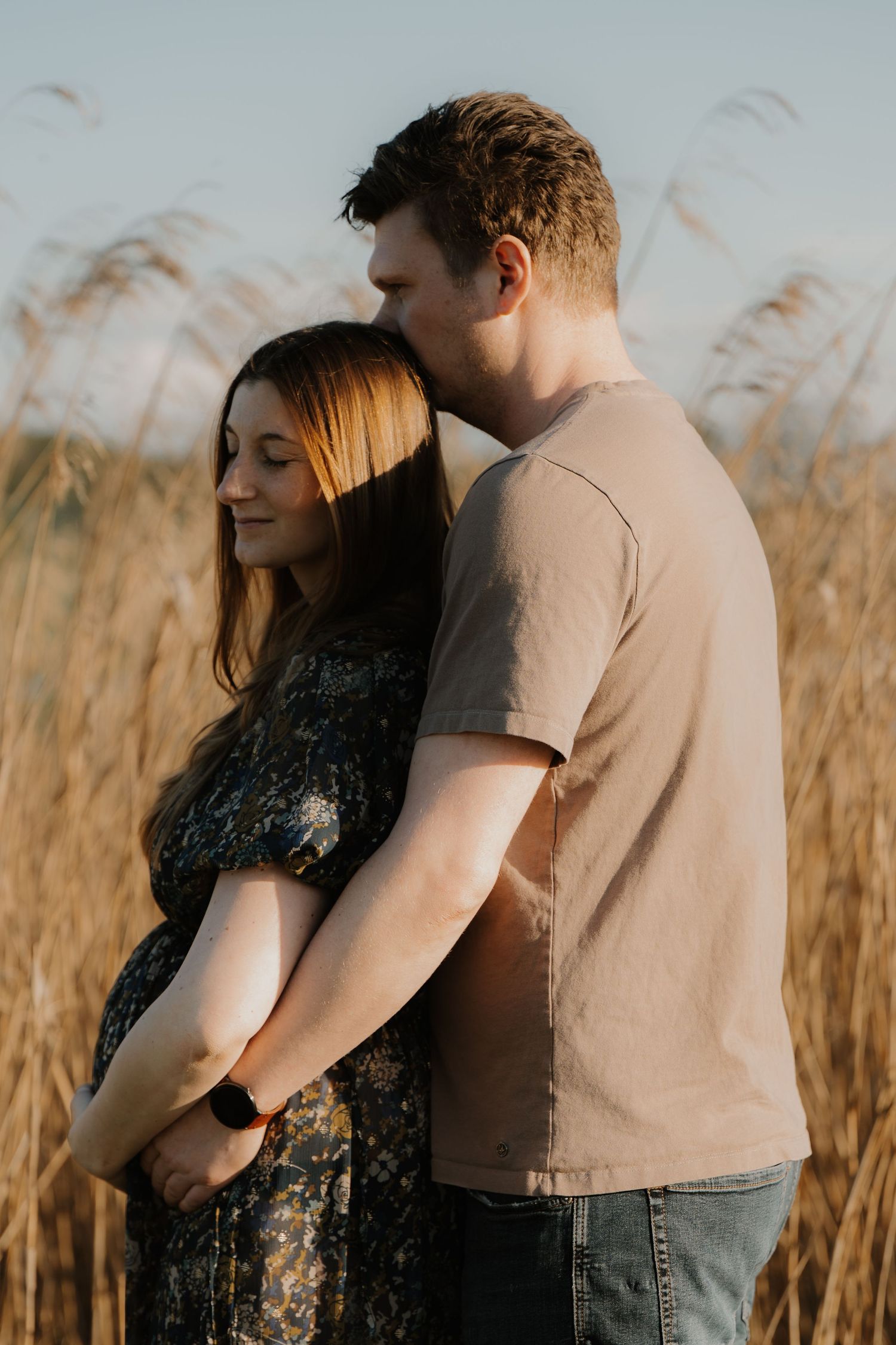 Couple embracing in golden field at sunset, pregnant woman in floral dress leaning back against partner.