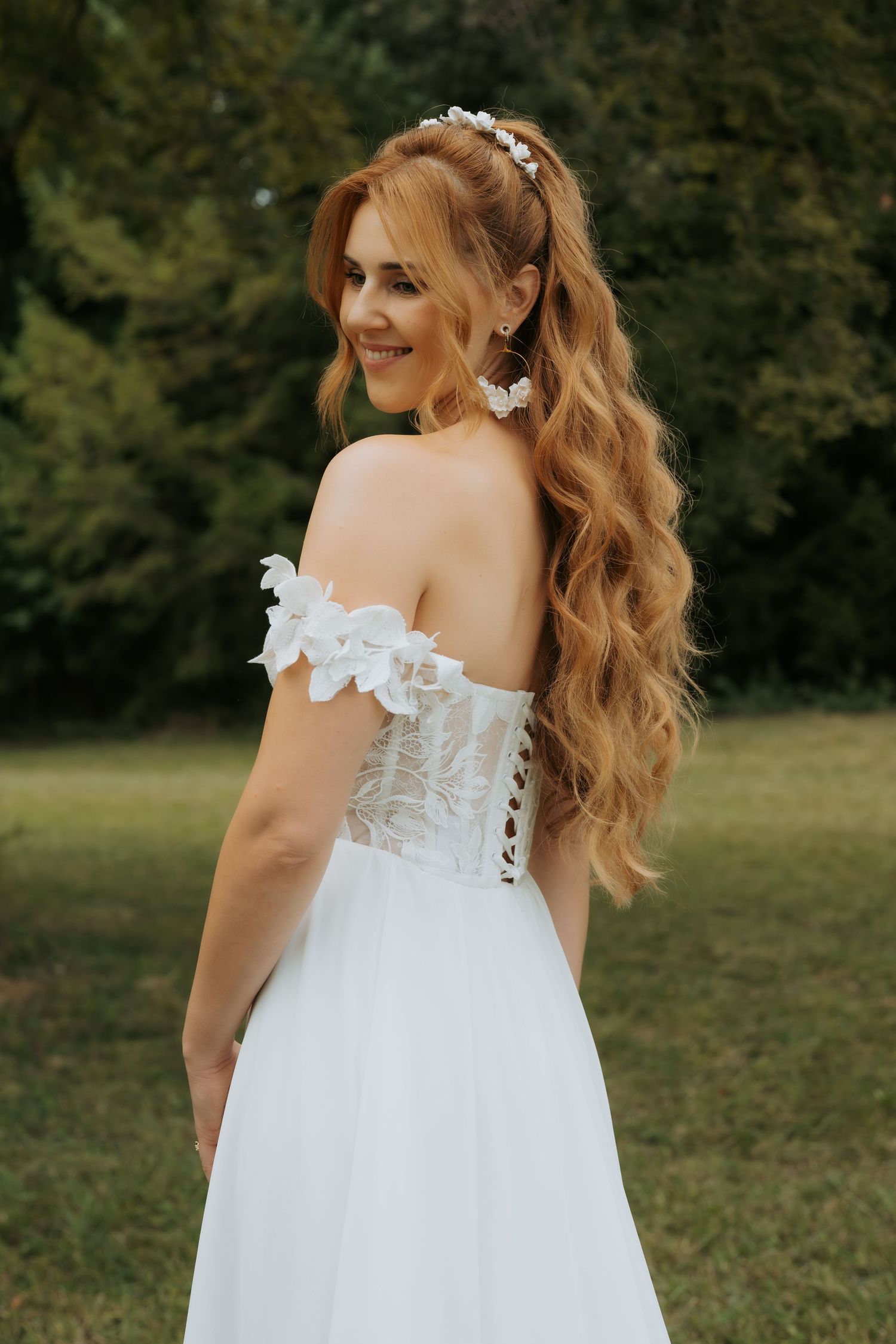 Smiling bride in off-shoulder lace wedding gown with floral headpiece and wavy auburn hair poses outdoors.