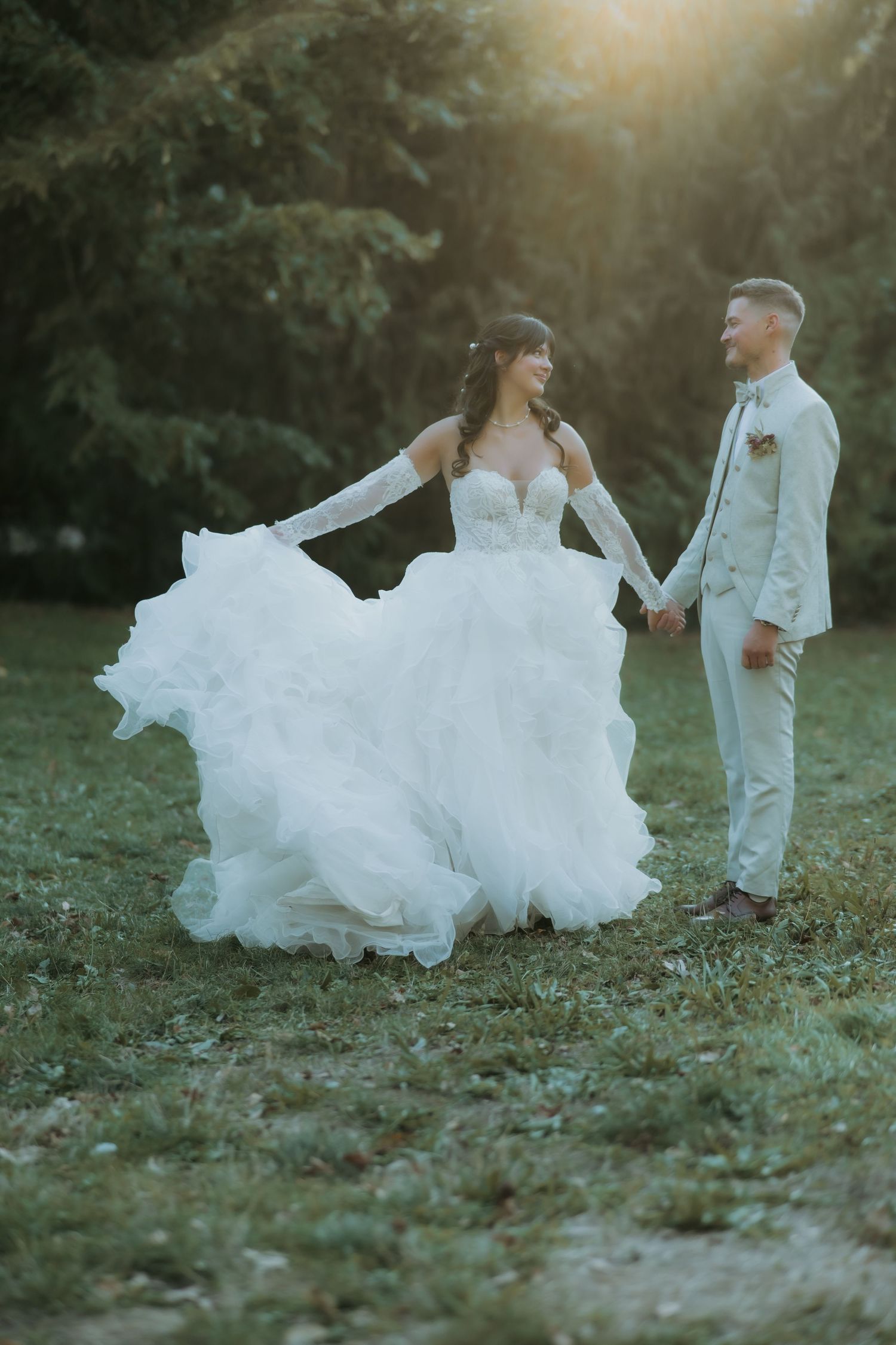 Bride in flowing white ballgown holds hands with groom in beige suit outdoors at golden hour.