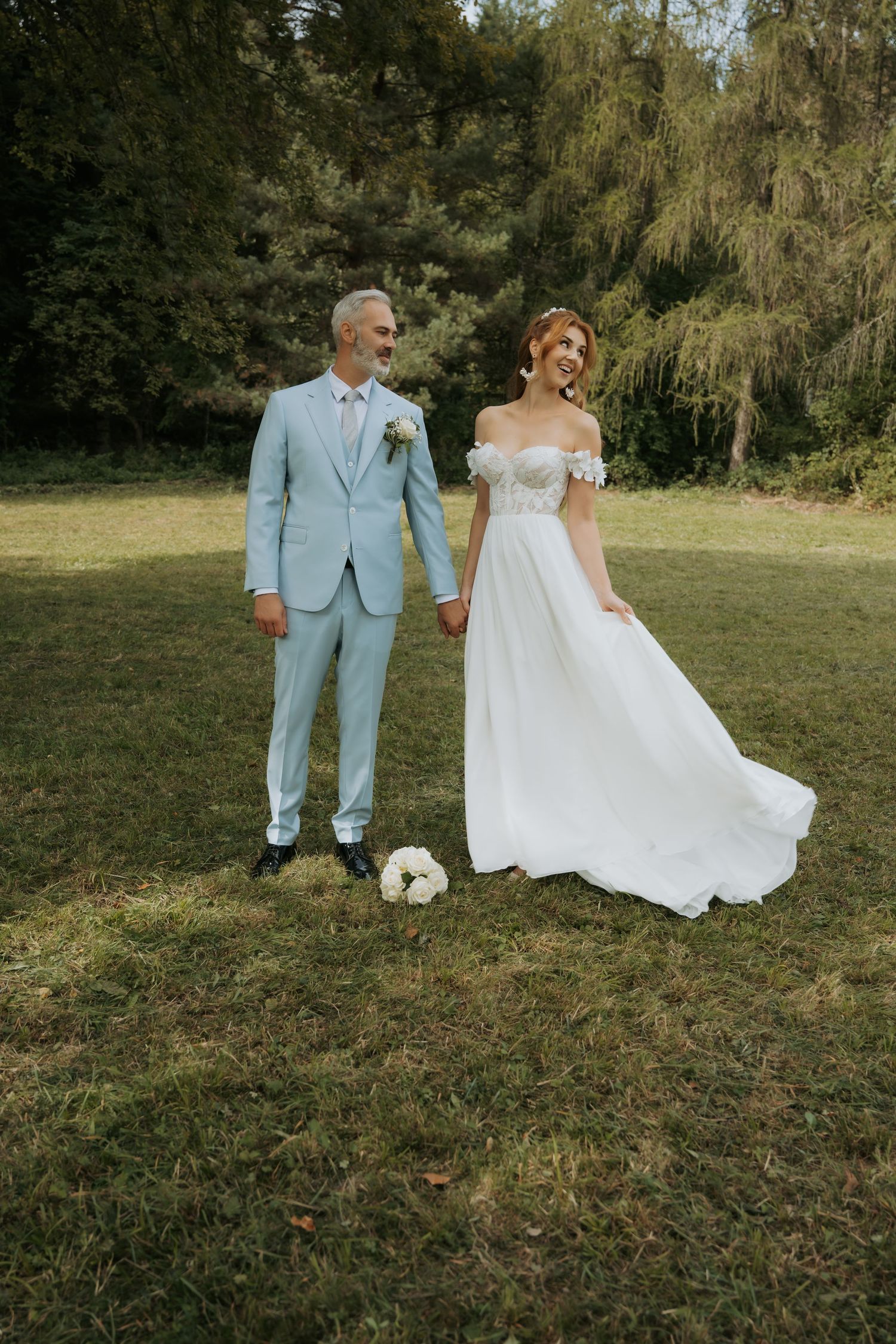 Bride in white off-shoulder gown and groom in light blue suit hold hands outdoors on green lawn.