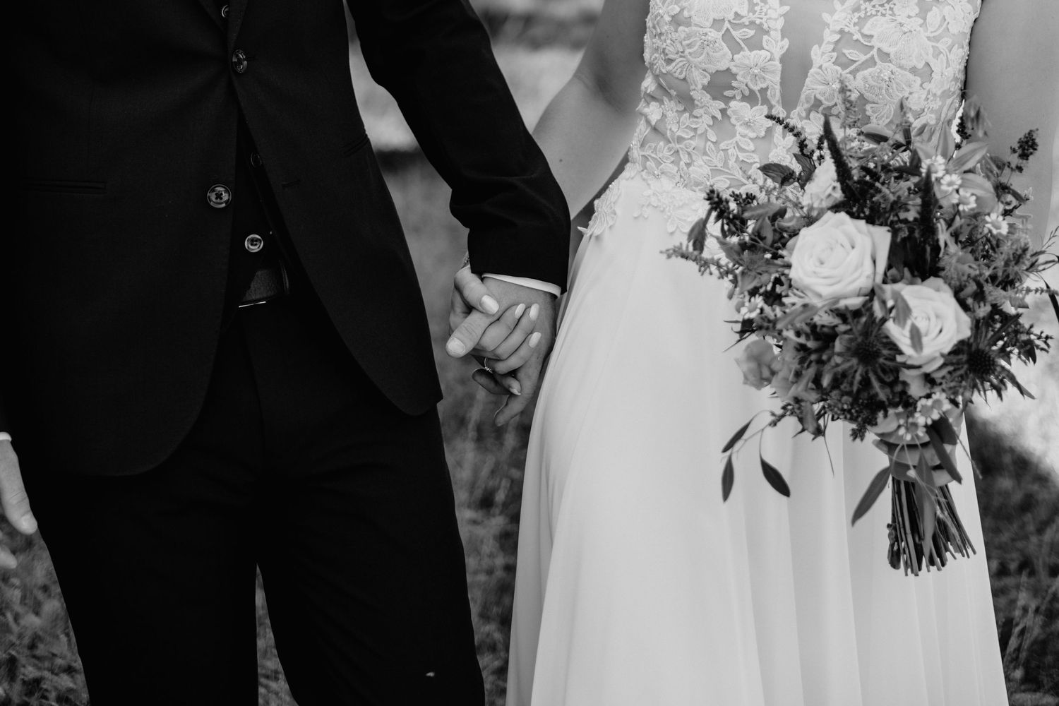Black and white photo of bride and groom holding hands, bride holding floral bouquet with roses and greenery.
