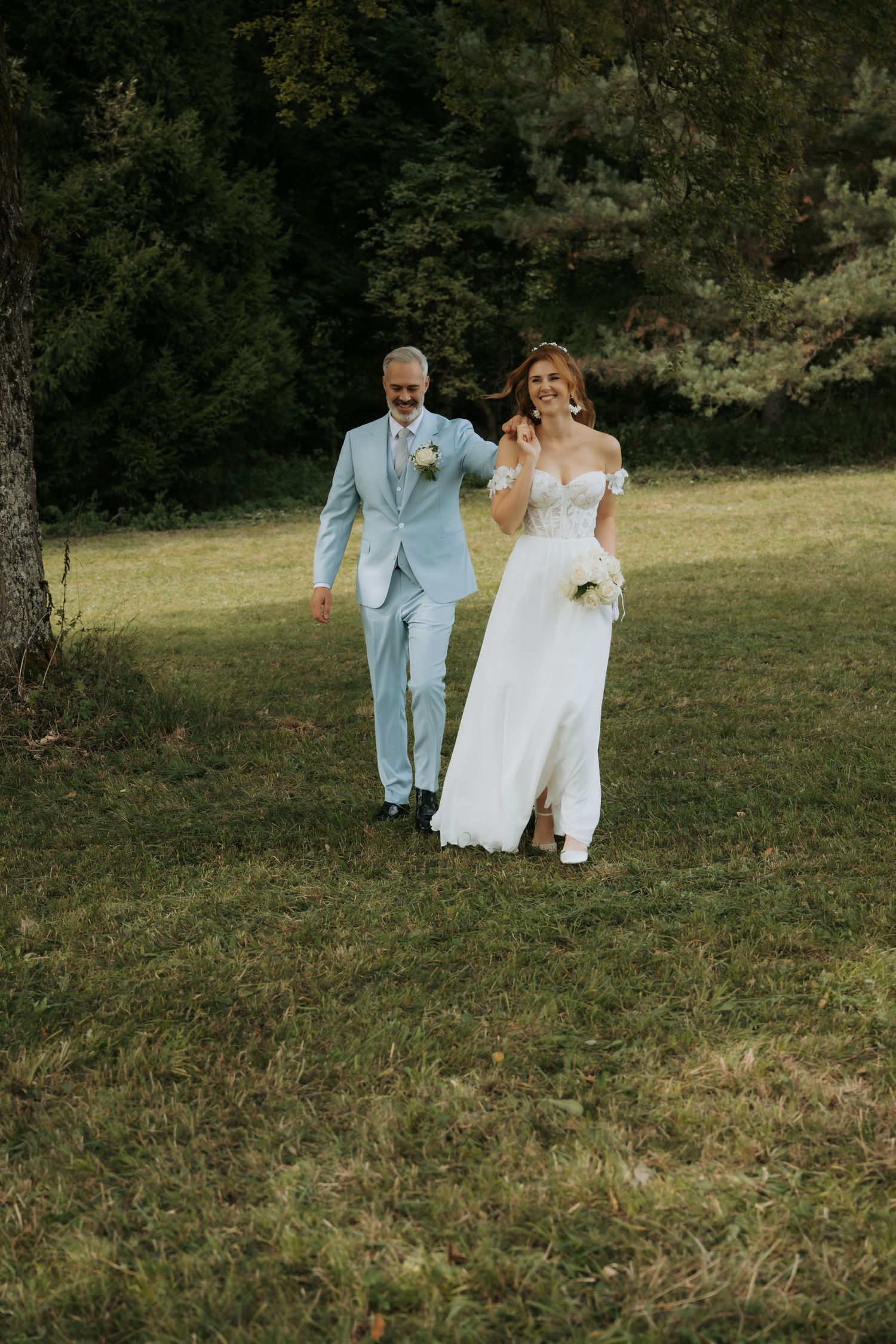 Bride and groom walking on green grass in a garden, bride in white lace gown holding bouquet, groom in light blue suit.