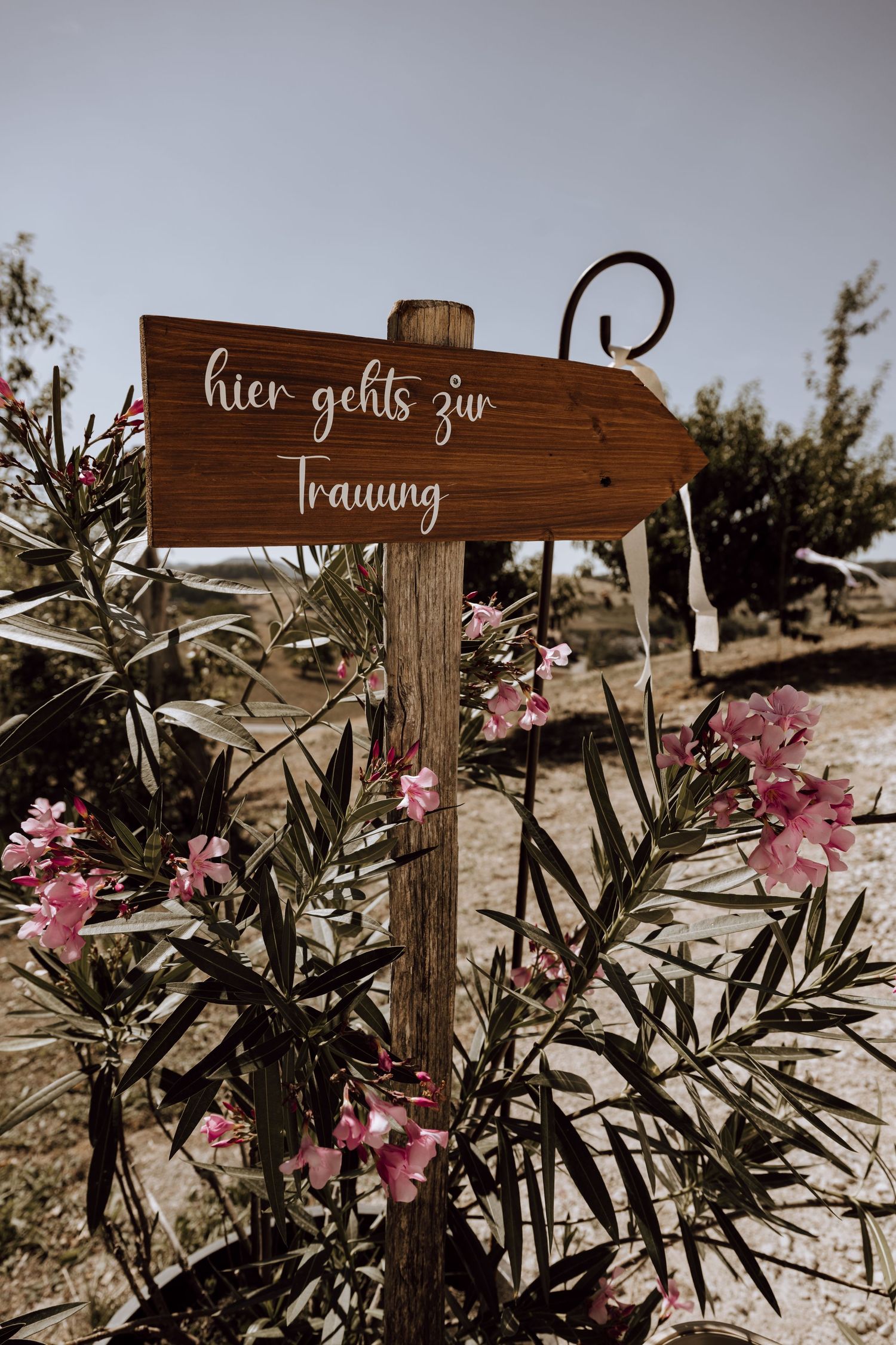 Wooden arrow sign reading 'hier gehts zur Trauung' surrounded by pink oleander flowers at an outdoor wedding.