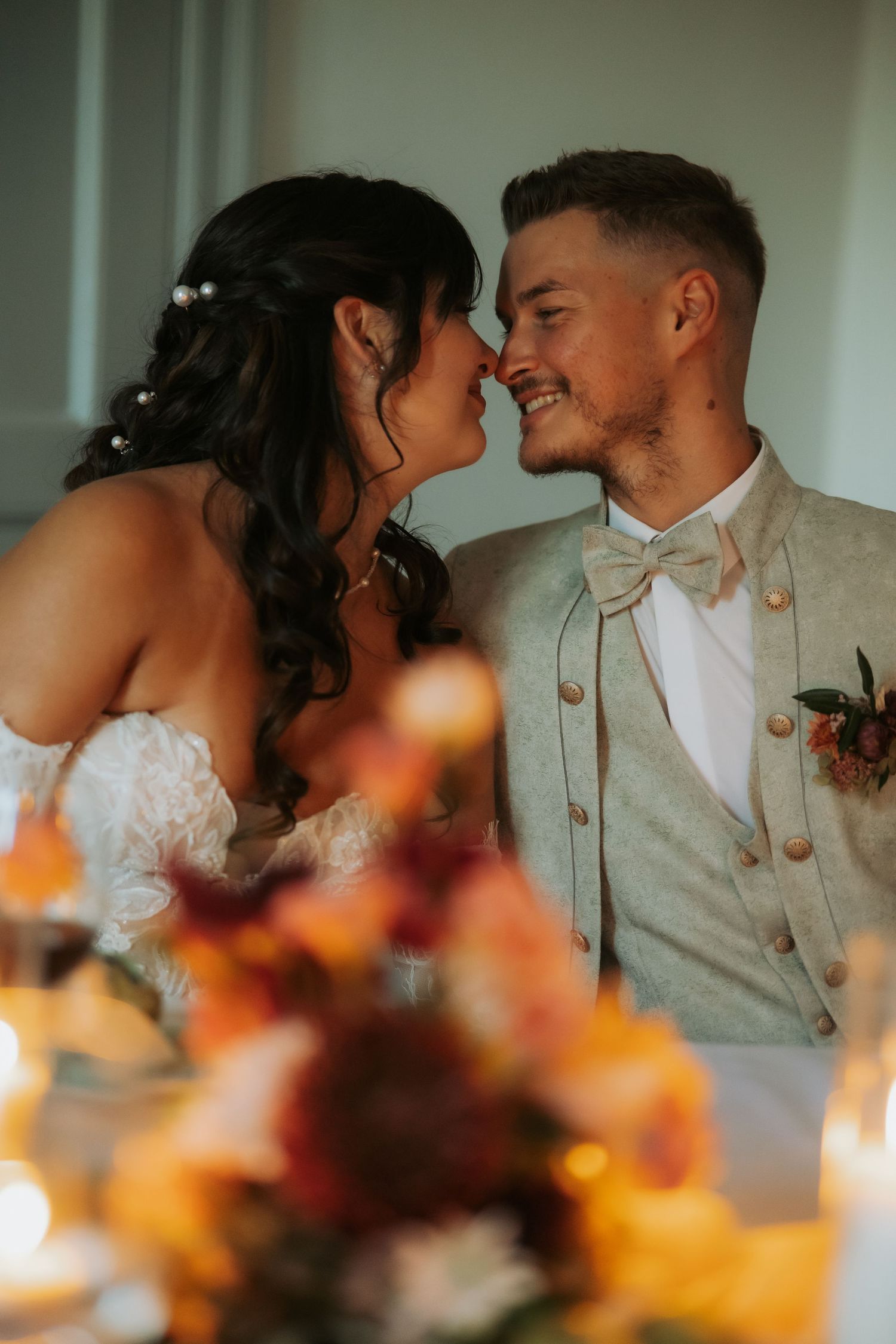 A smiling groom in a sage suit leans in to kiss his bride in a white dress at their romantic candlelit wedding reception.