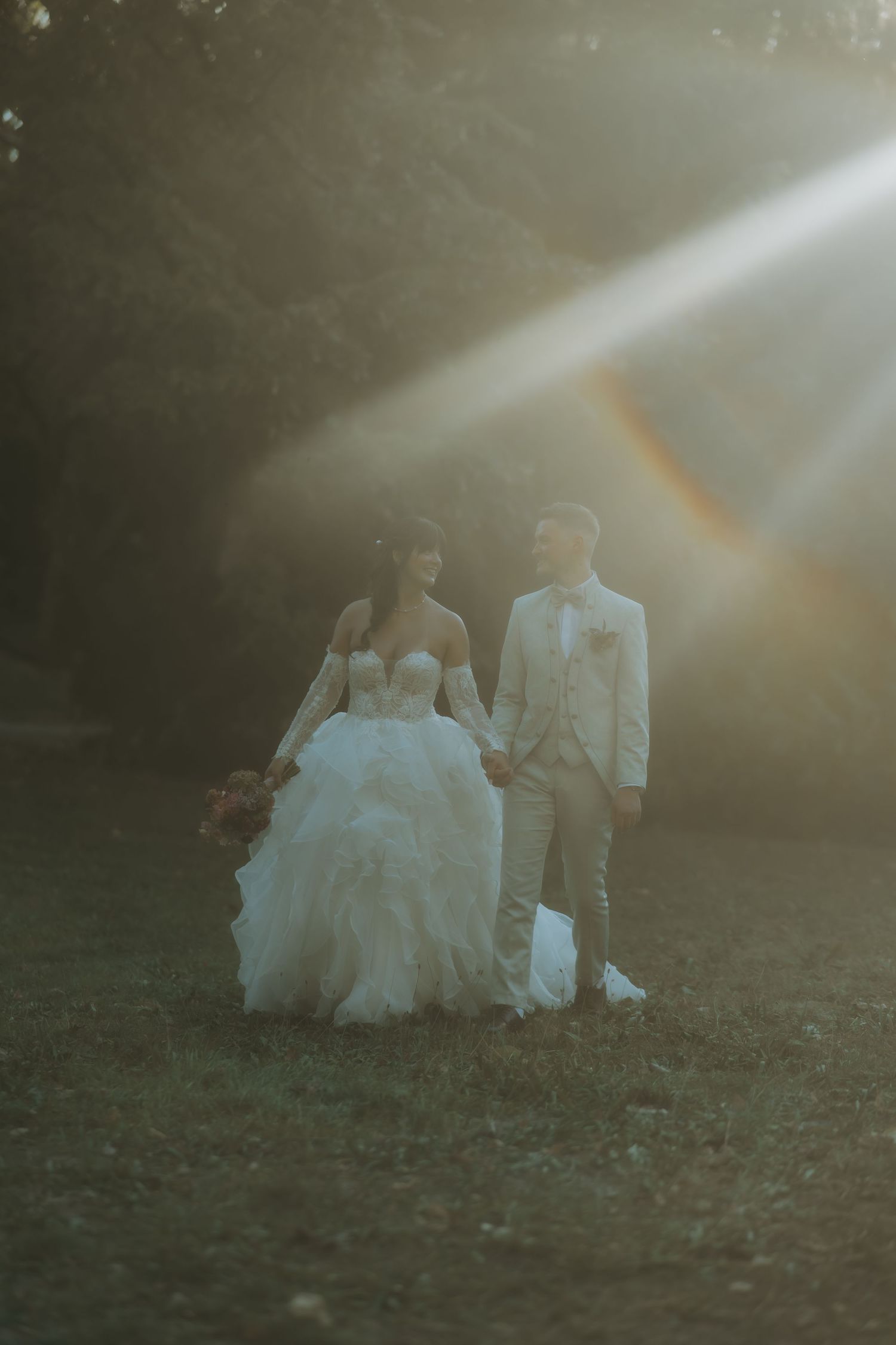 Bride and groom hold hands walking in golden sunlight rays in a lush garden, bride wearing a ruffled white ball gown.