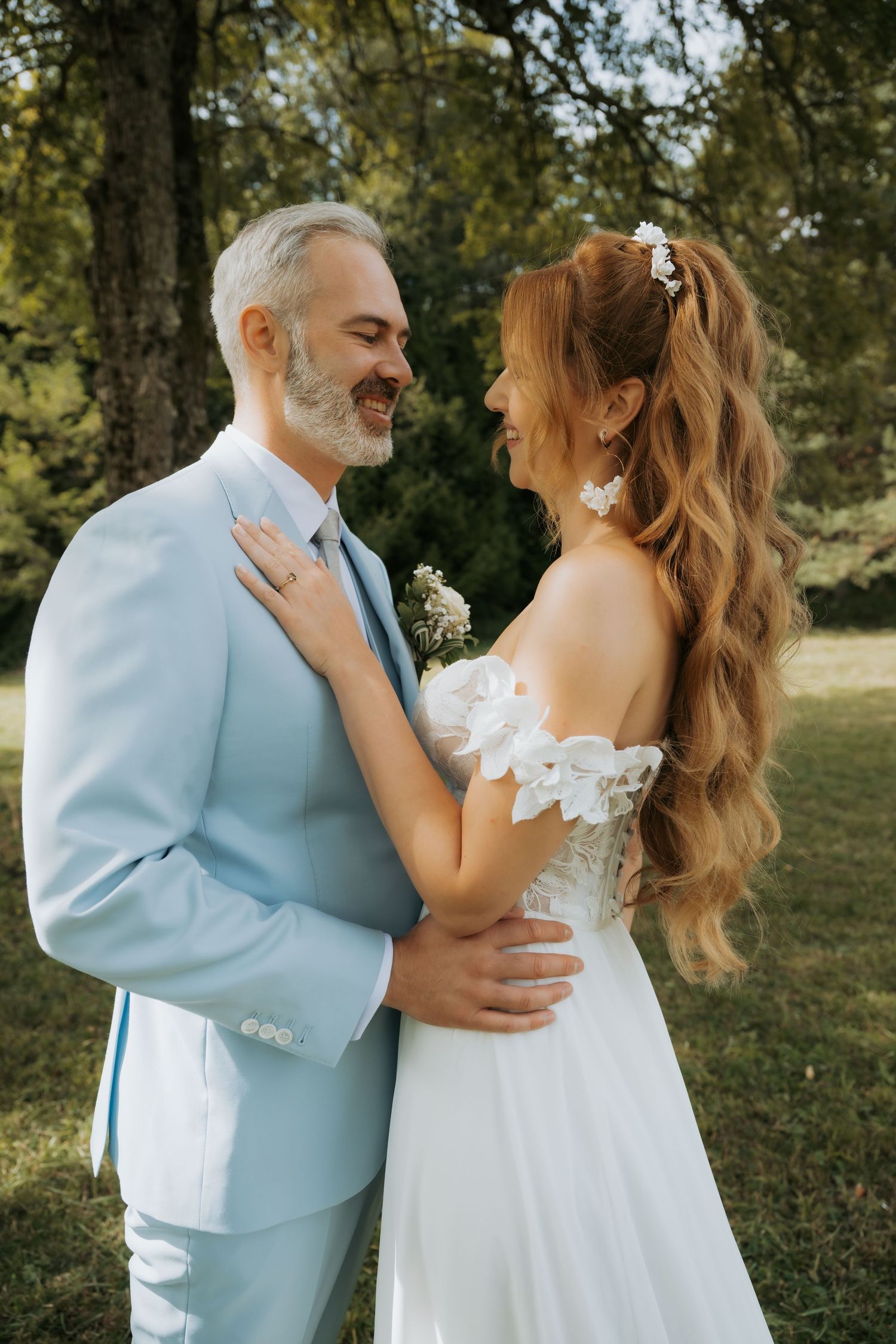 A smiling bride and groom in a garden, she wears a white lace gown, he wears a light blue suit.