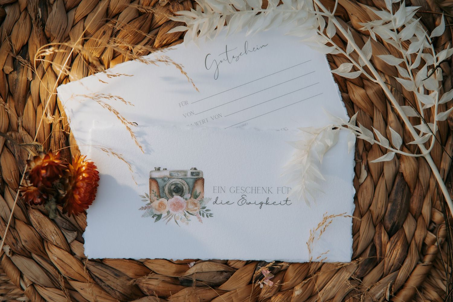 White gift card envelope with floral camera design on woven basket with dried flowers and feathers.