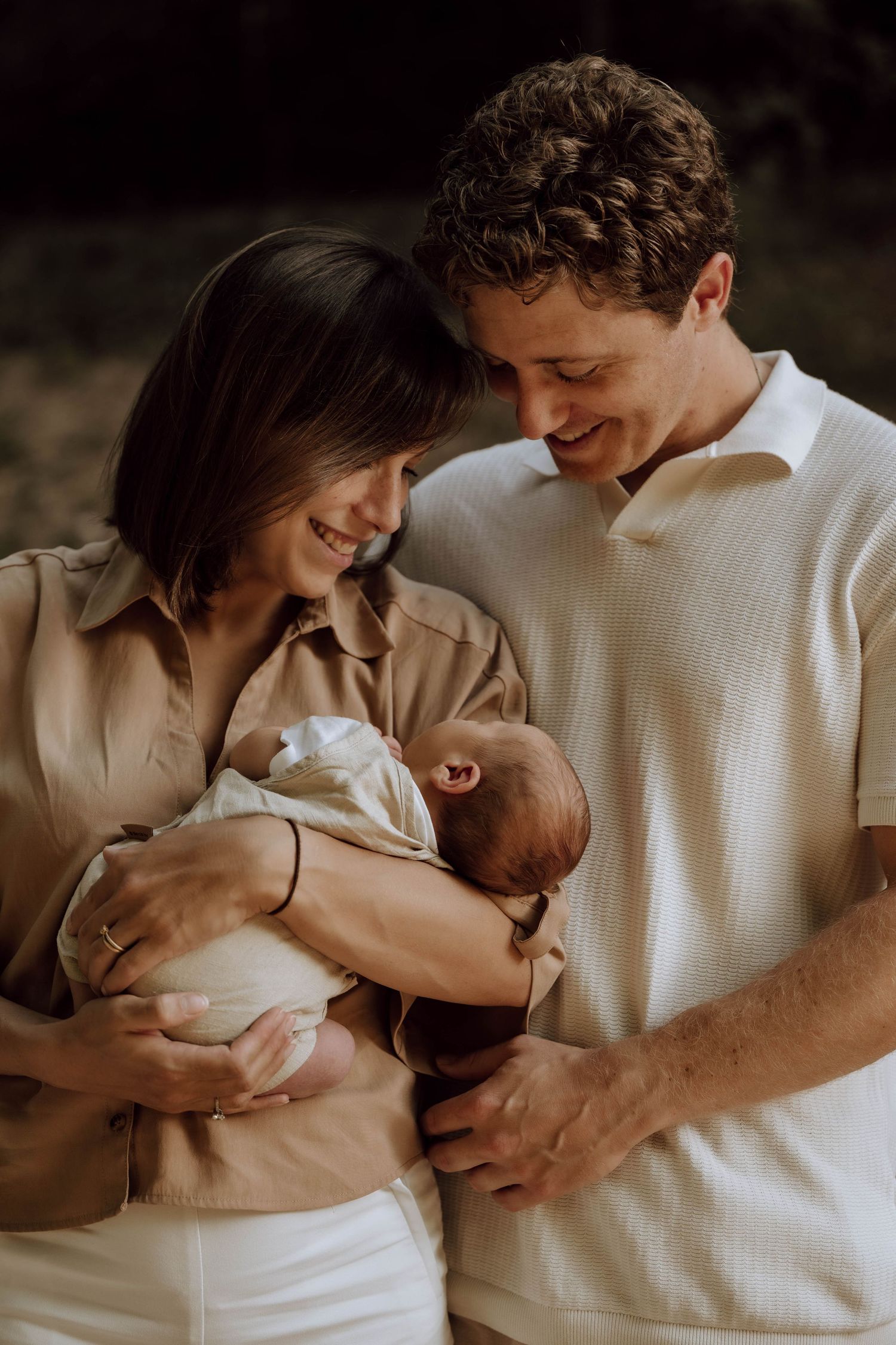 Happy couple smiling down at their newborn baby during an outdoor family portrait session.