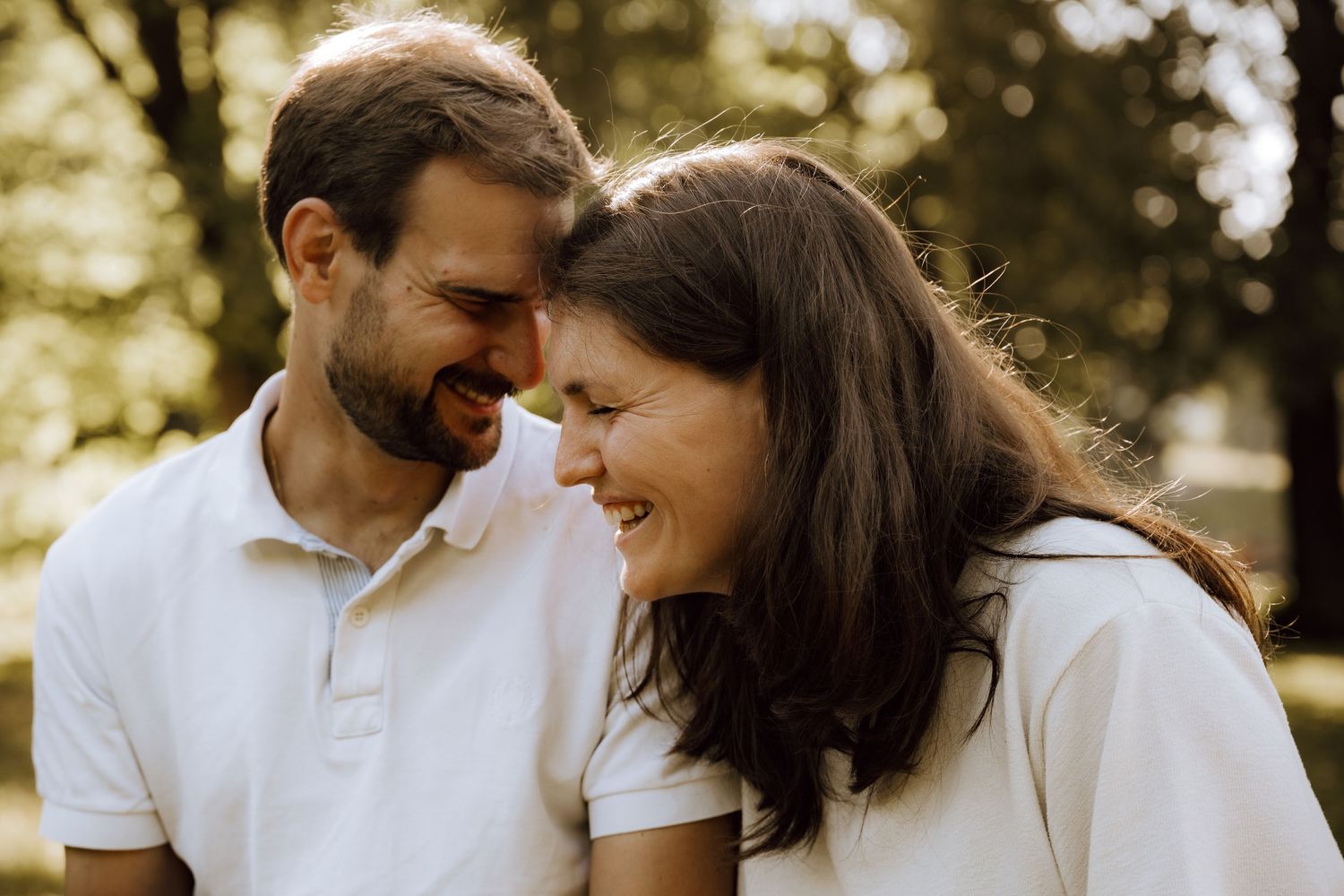 A happy couple in white shirts share a joyful moment outdoors, touching foreheads and smiling in a sunny park.