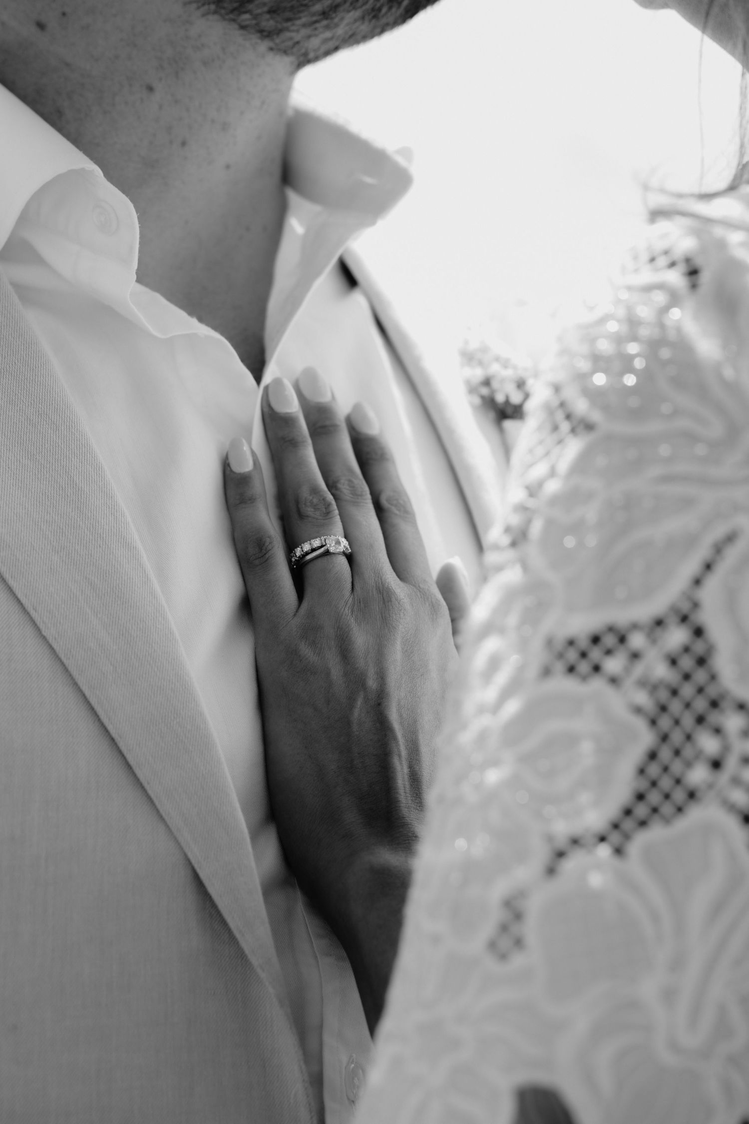 Black and white photo of bride placing hand on groom's chest, showcasing elegant wedding ring and lace dress detail.