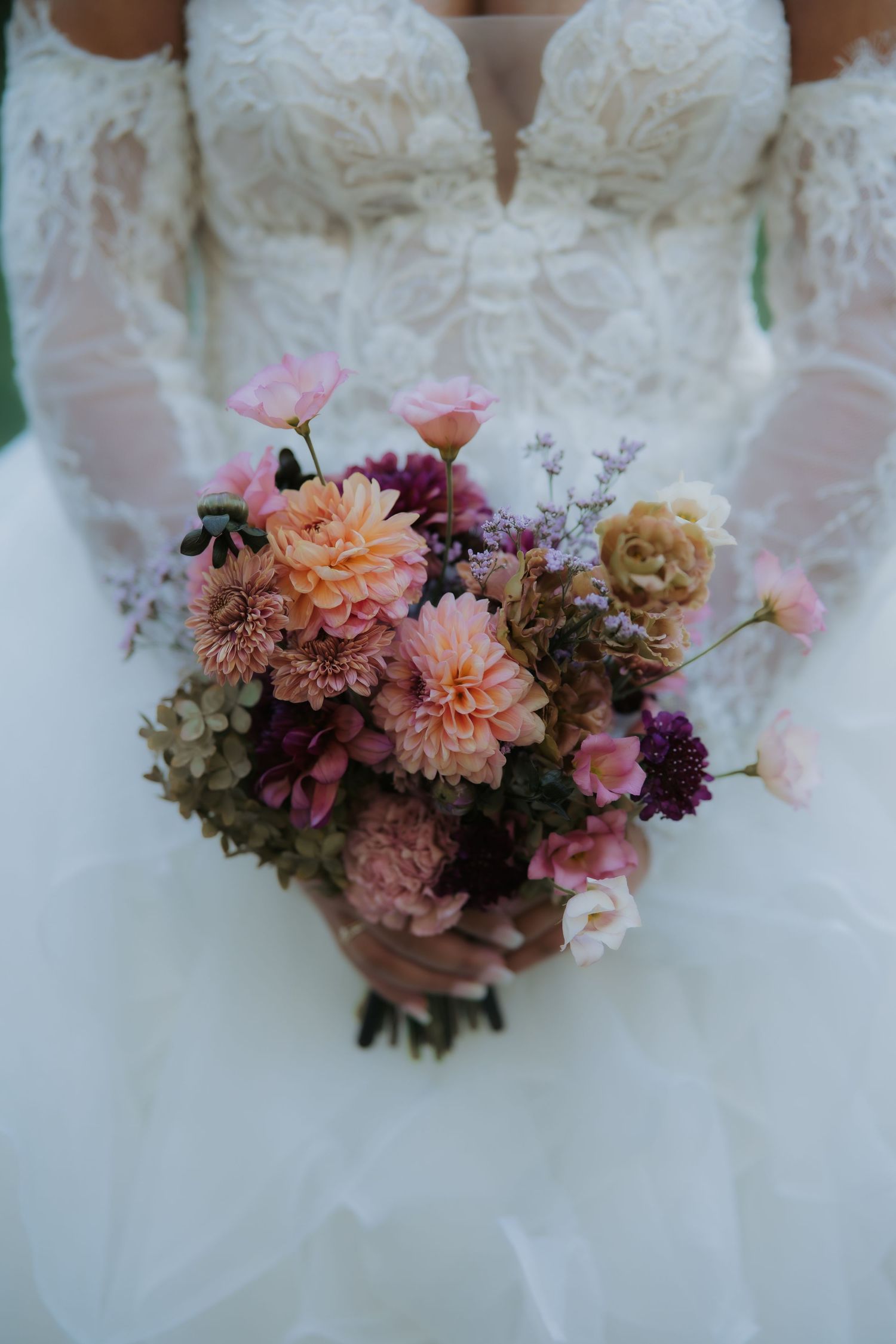 Bride holding a lush wedding bouquet with pink, peach, and burgundy dahlias in a lace long-sleeve gown.