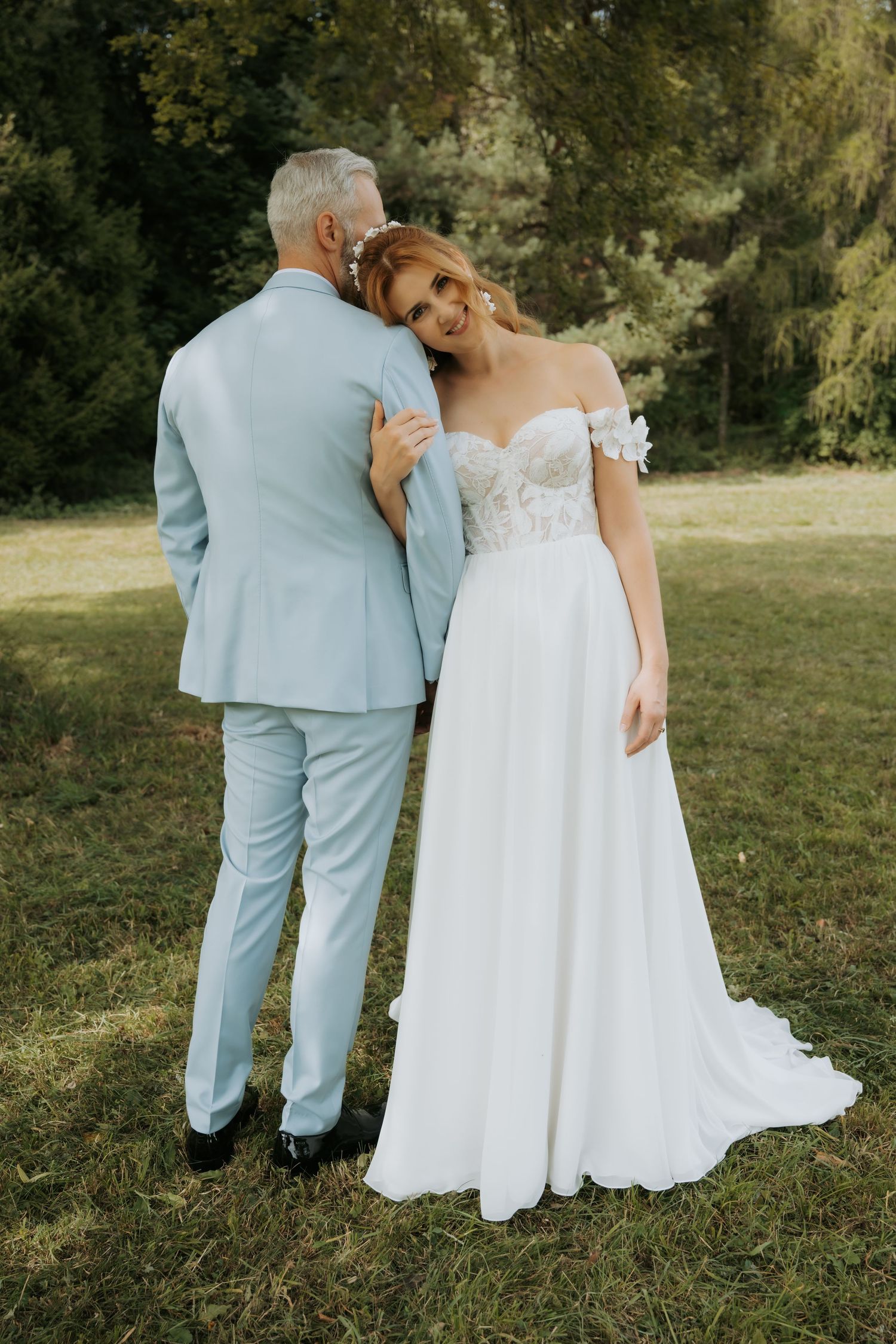A bride in a white off-shoulder gown embraces a groom in a light blue suit outdoors on green grass.