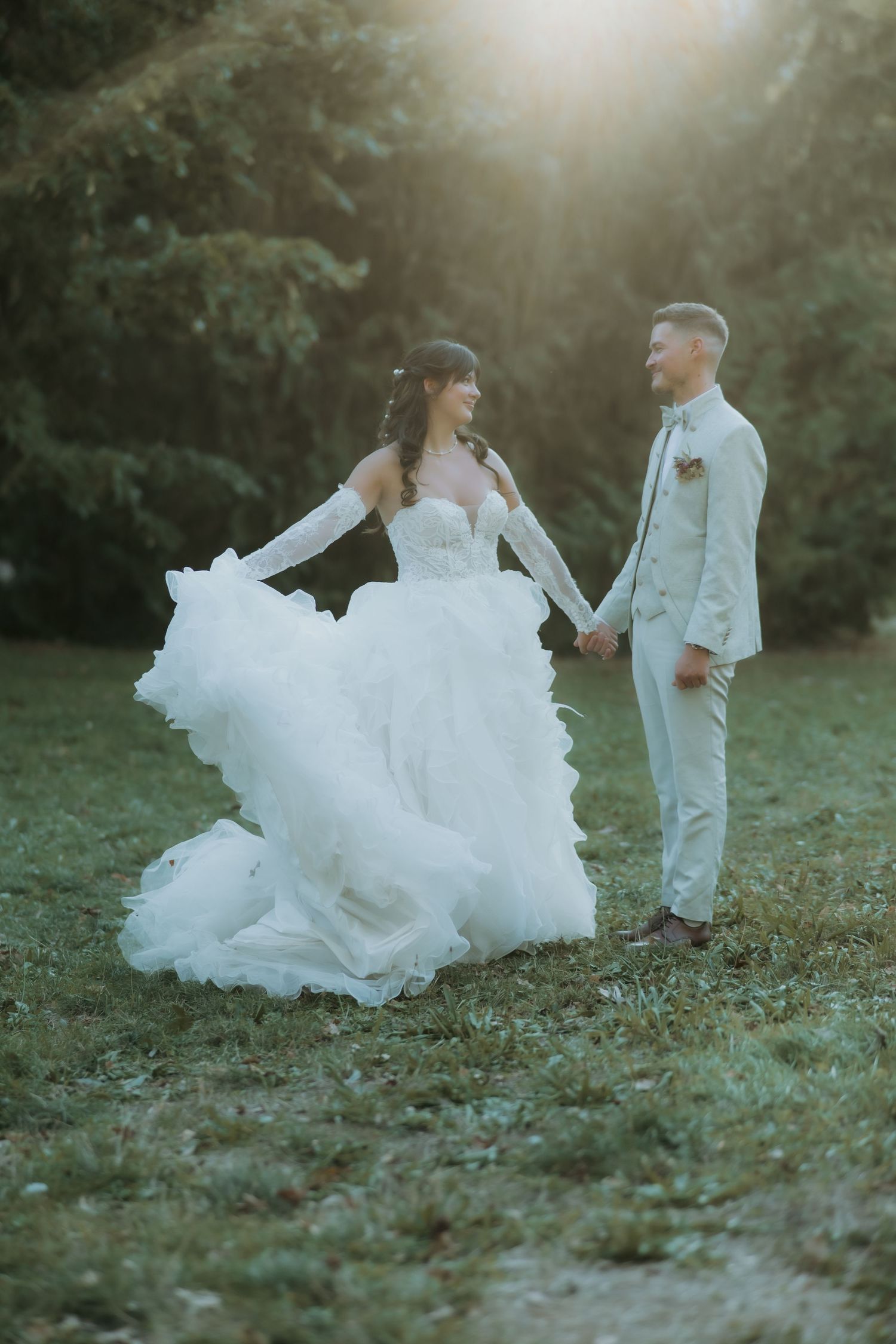 Bride twirls her flowing white ruffled wedding gown while holding grooms hand in a sunlit green garden.