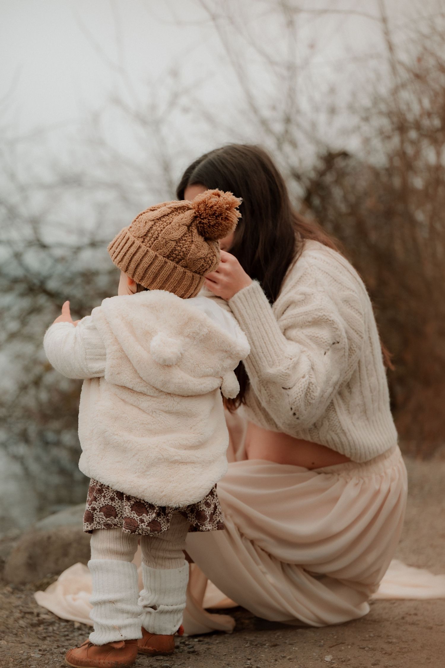 A mother crouches down to embrace her toddler wearing a brown pom-pom hat and cream jacket outdoors.