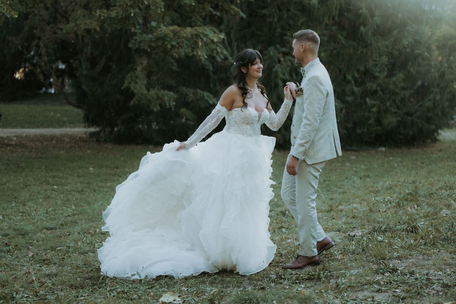 A bride in a flowing white gown and groom in a beige suit hold hands while dancing outdoors in a lush green park.