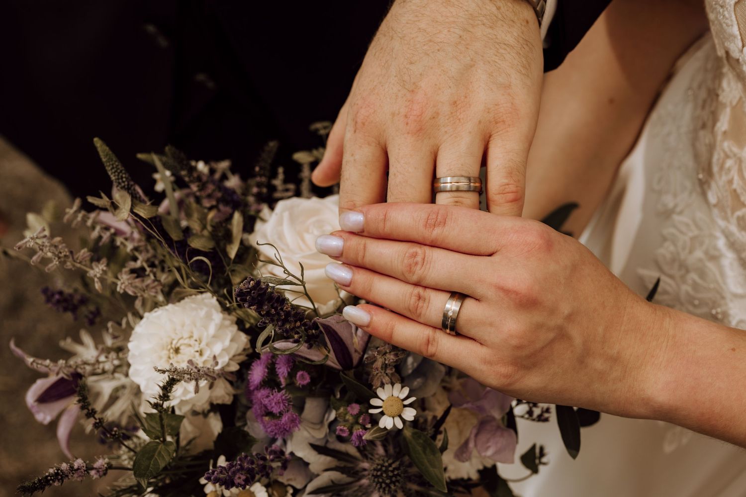 Newlyweds displaying wedding rings on hands resting over a purple and white floral bouquet.