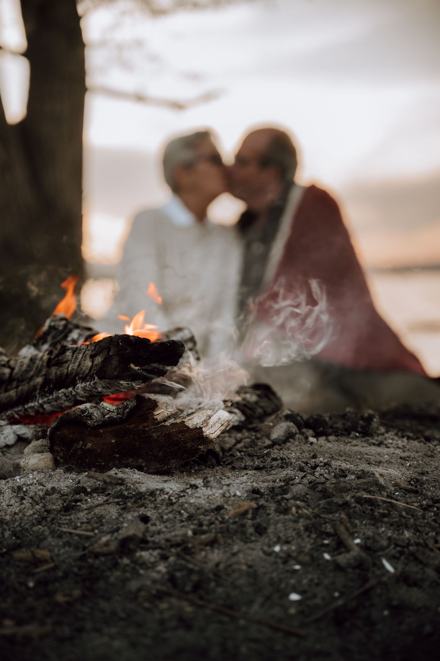 Couple kissing by a glowing campfire outdoors at dusk, wrapped in a blanket near a tree.