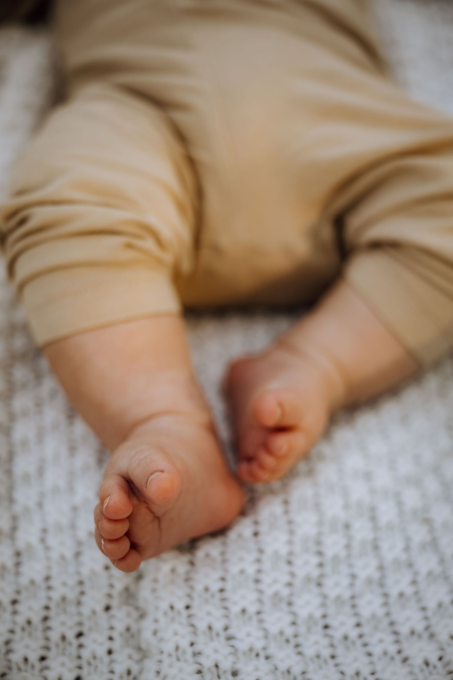 Close-up of tiny newborn baby feet in beige pajamas resting on a white knit blanket.