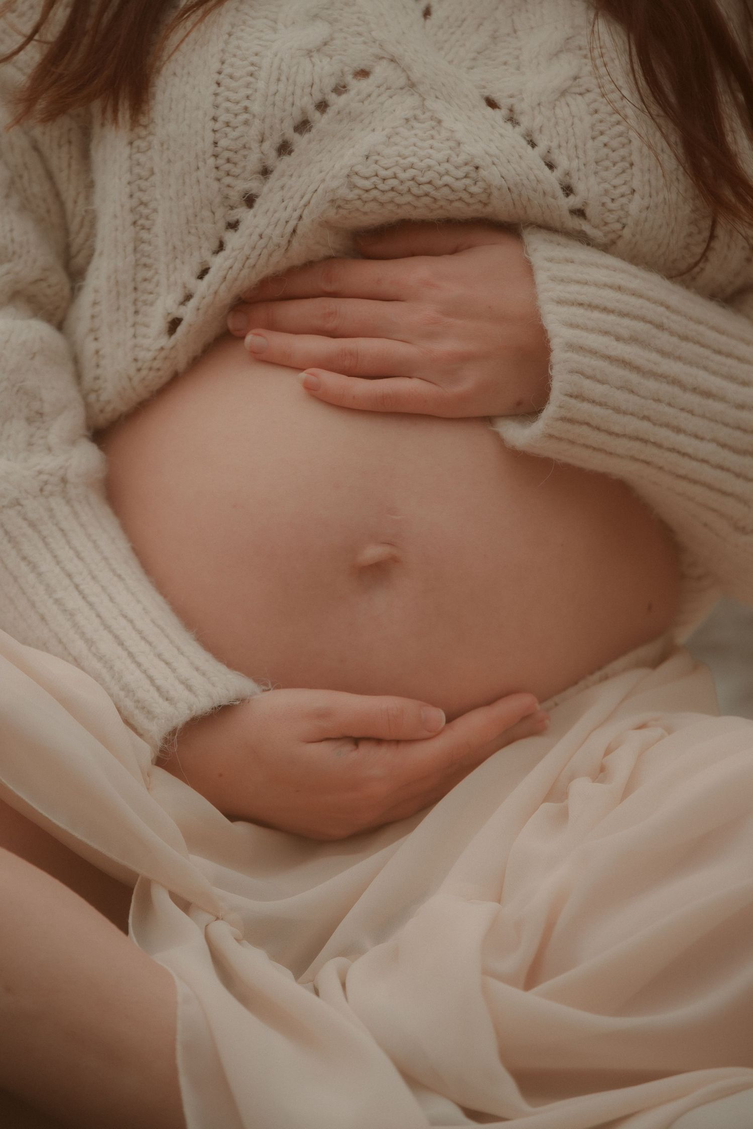 Close-up of a pregnant woman in a cream knit sweater gently cradling her bare baby bump.