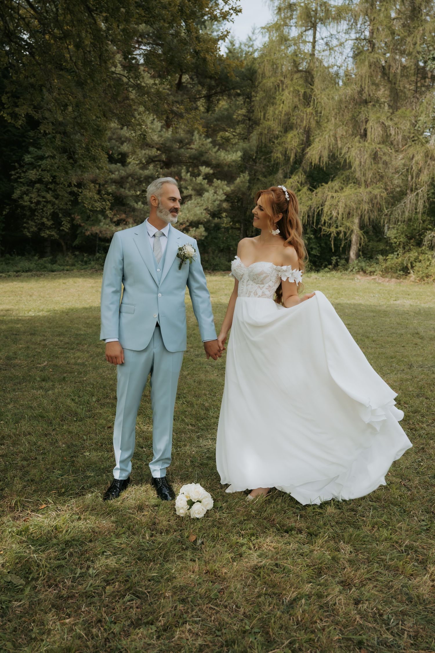 A bride in a white off-shoulder gown and groom in a light blue suit hold hands on a green lawn with trees behind.