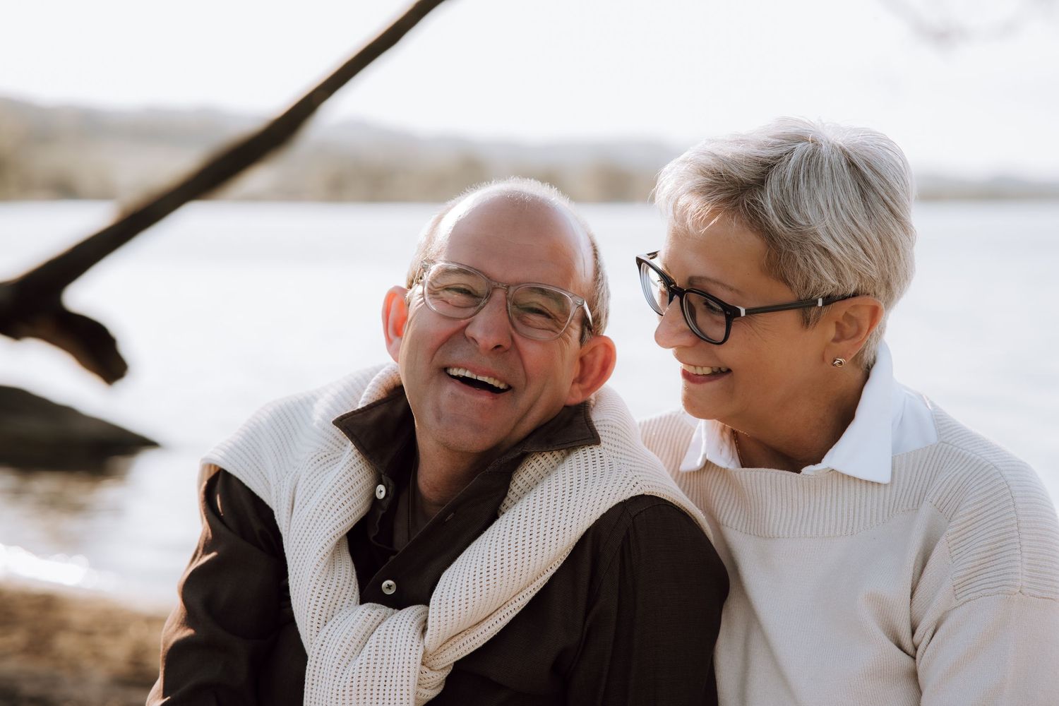 Happy elderly couple laughing together outdoors near a lake, enjoying a warm moment in casual winter attire.