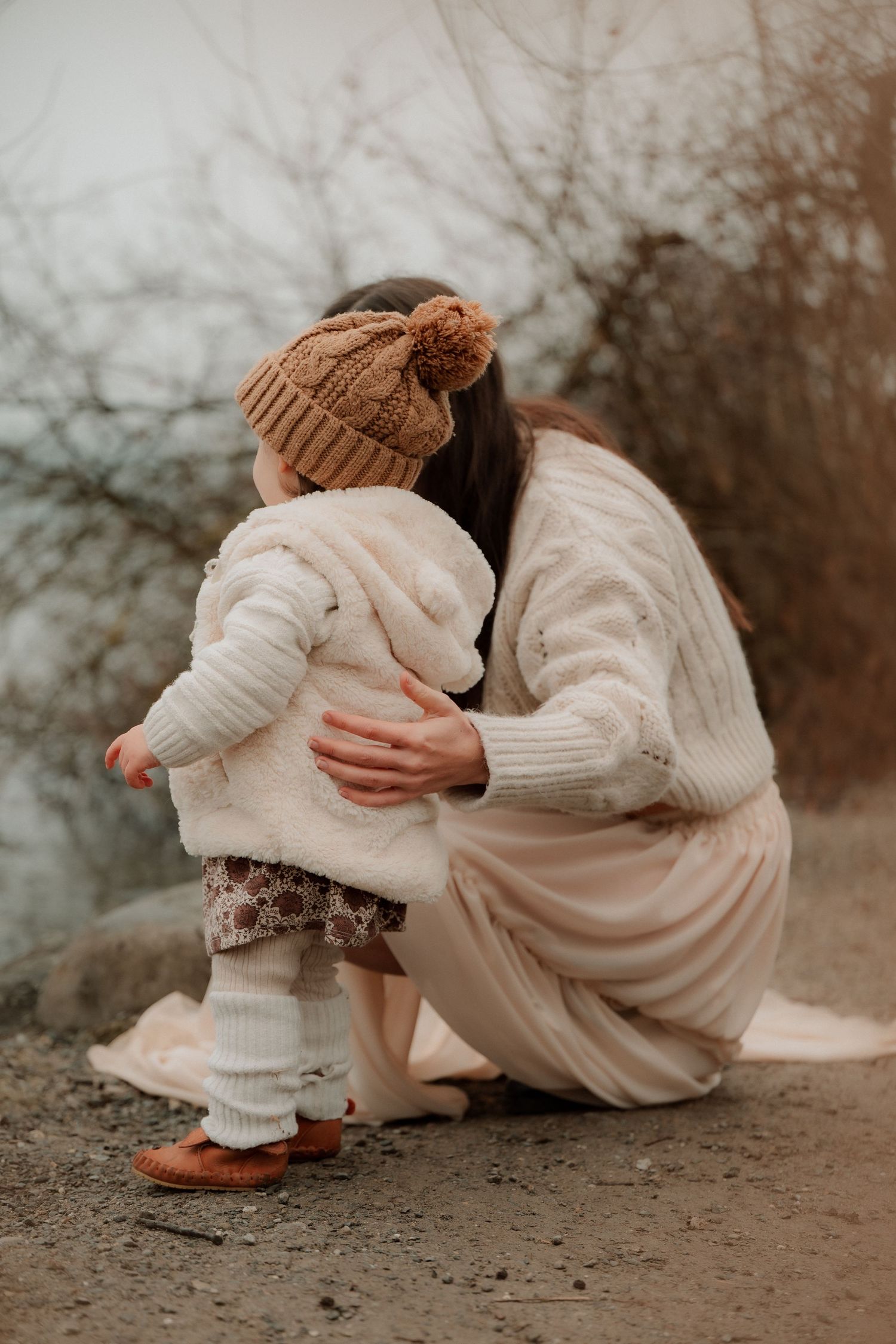 Mother in cream sweater crouches to embrace toddler in knit hat outdoors in autumn setting.