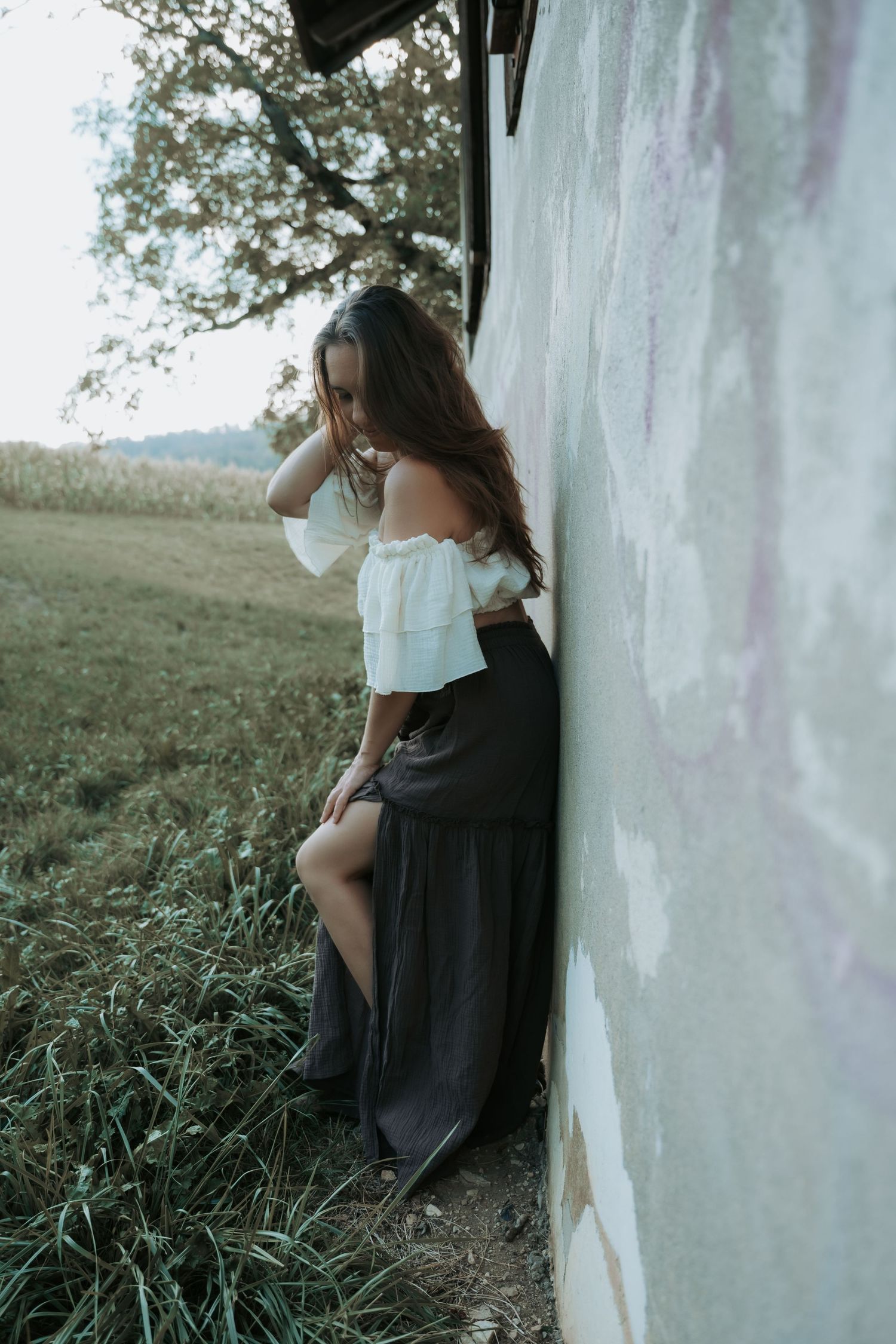 Young woman in white off-shoulder top and dark maxi skirt leaning against rustic wall in countryside setting.