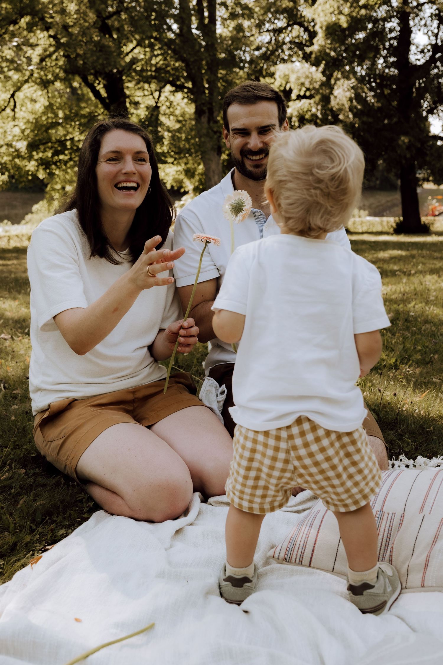 A joyful family enjoys a park picnic as a toddler presents dandelion flowers to his laughing mother.
