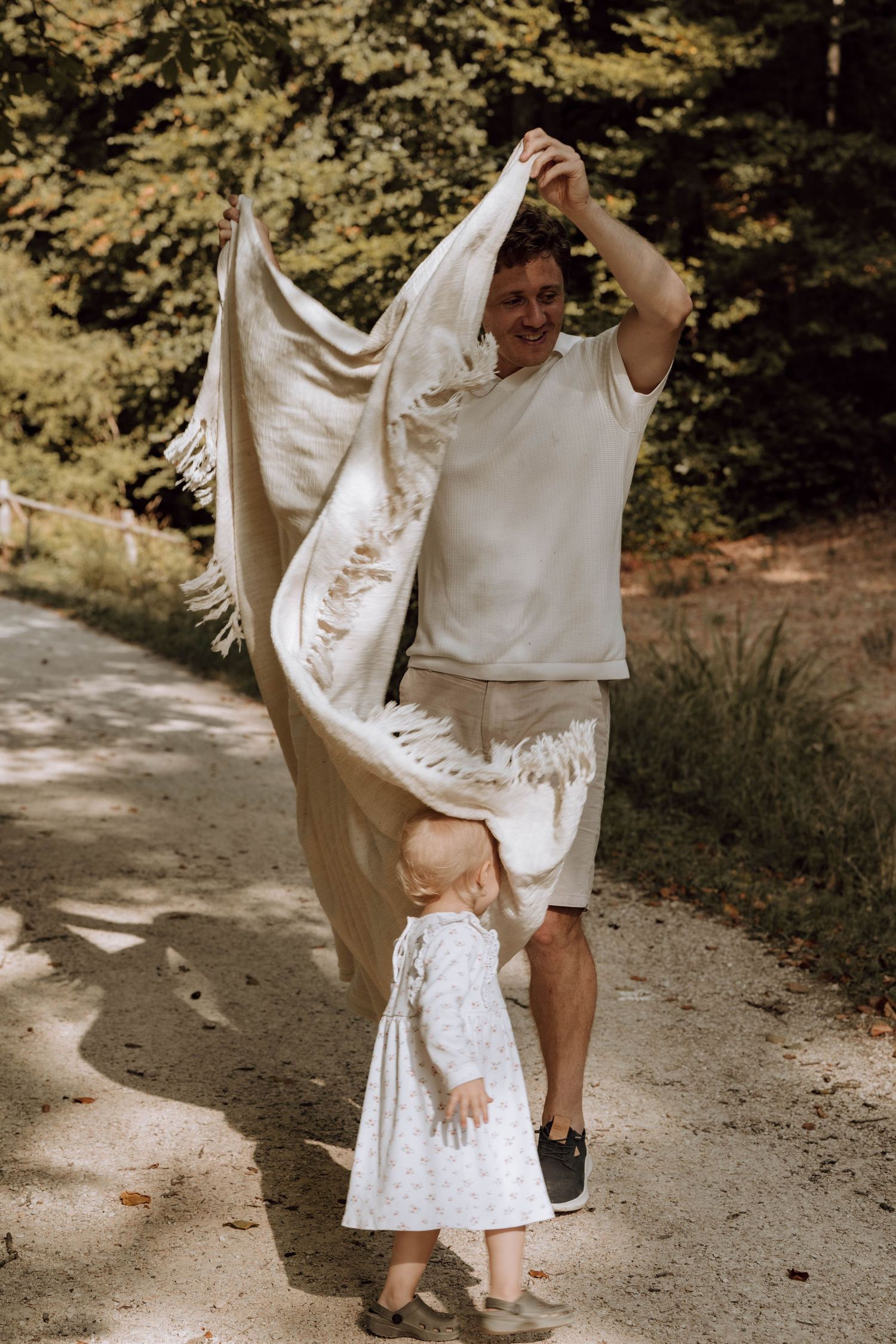 Man playfully waving a fringe blanket over a toddler girl on a sunny forest path.