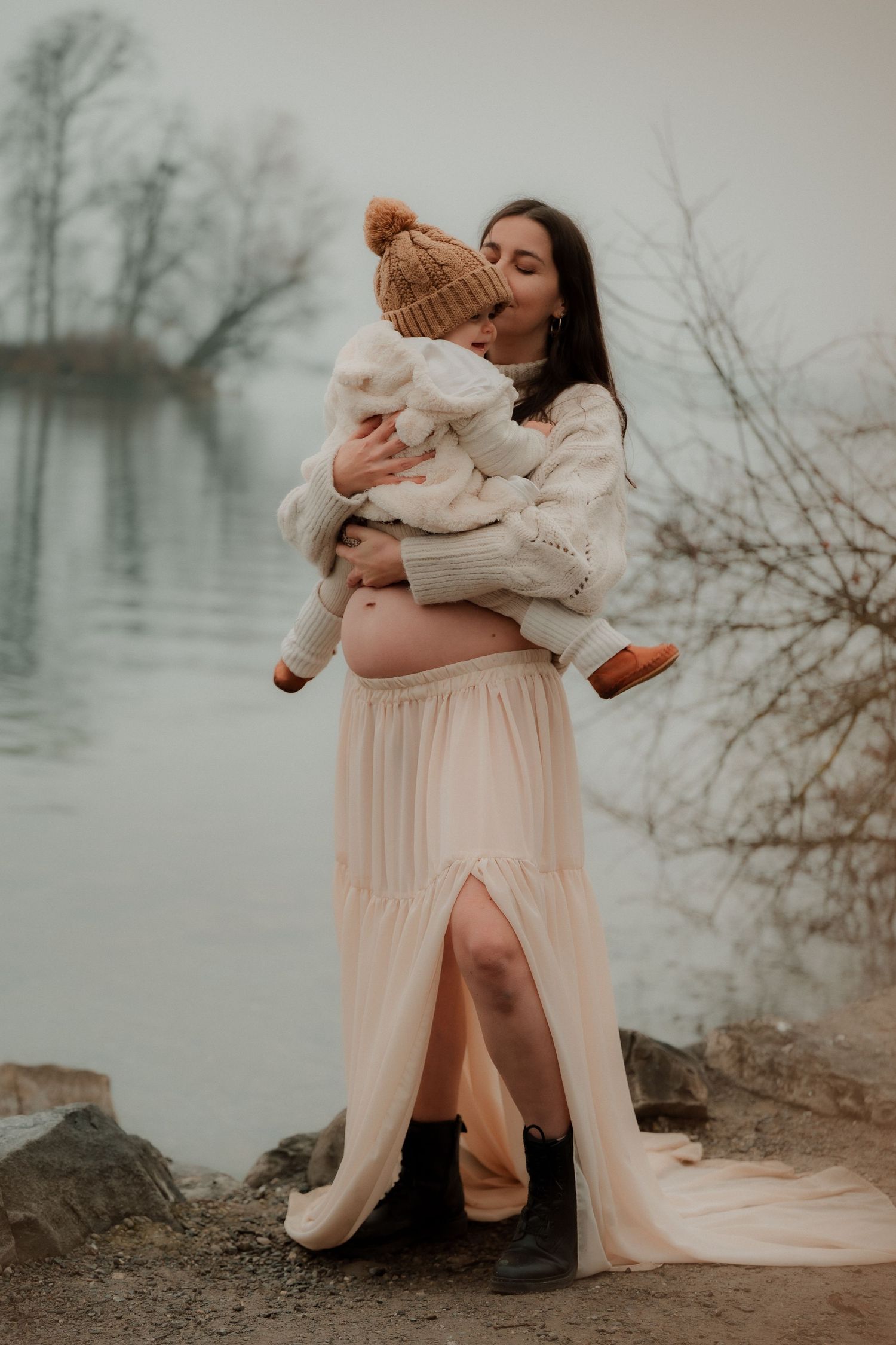 Pregnant woman in cream tulle skirt holds toddler in knit hat by misty lake in winter.