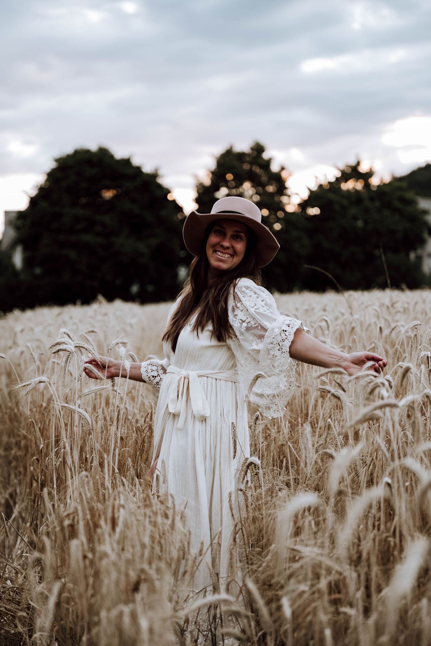Smiling woman in white lace dress and hat standing in golden wheat field with trees in background.