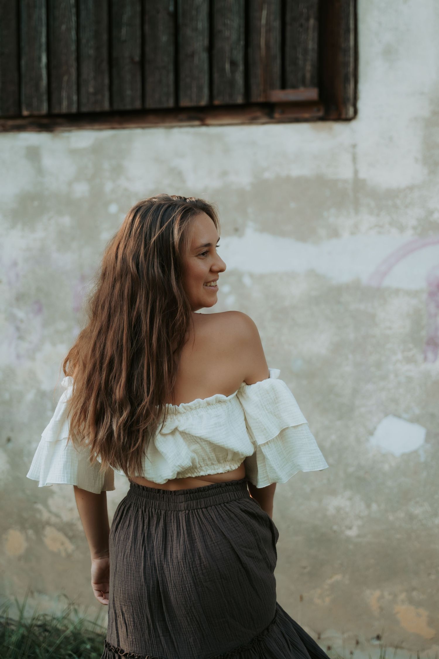 Young woman with long brown hair wearing white off-shoulder crop top and dark maxi skirt, smiling outdoors.