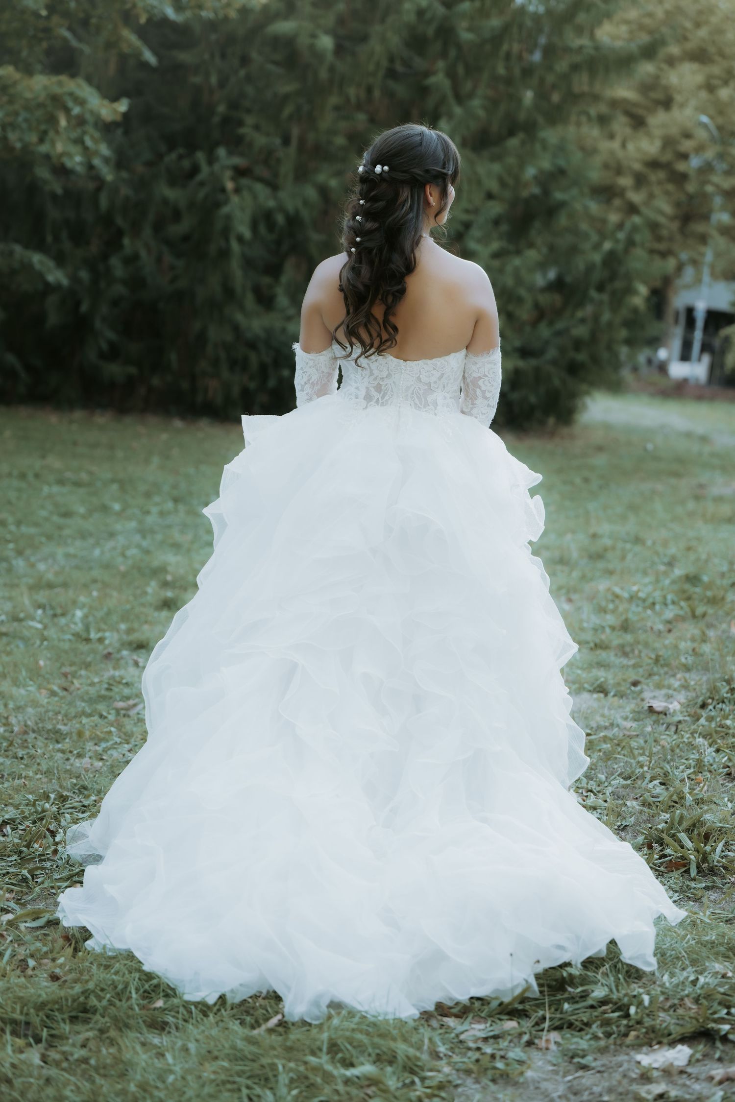 Bride in white ruffled off-shoulder wedding gown with lace gloves standing in garden, back view.